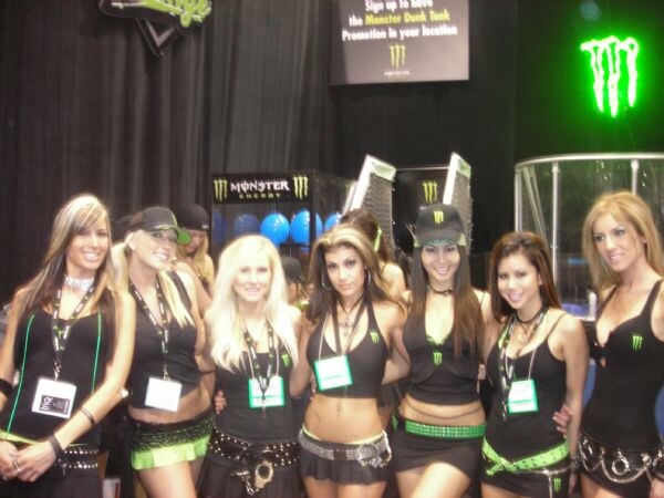 Seven women in black and green Monster Energy outfits, several wearing badges, stand in a row in front of a Monster Dunk Tank sign and a glowing green Monster logo.
