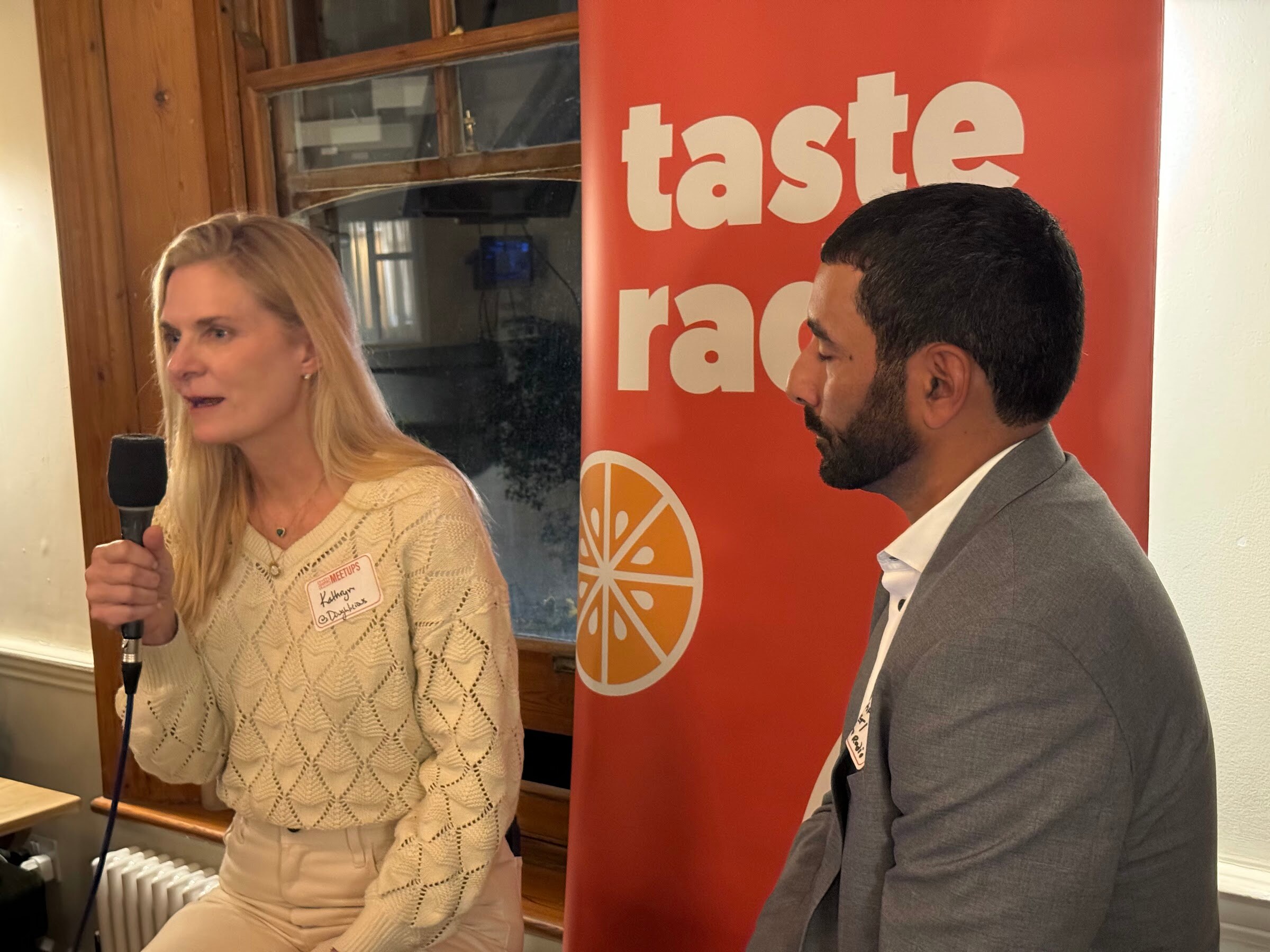 Blonde woman in a cream sweater holding a microphone beside the Taste Radio host, with a Taste Radio banner behind.