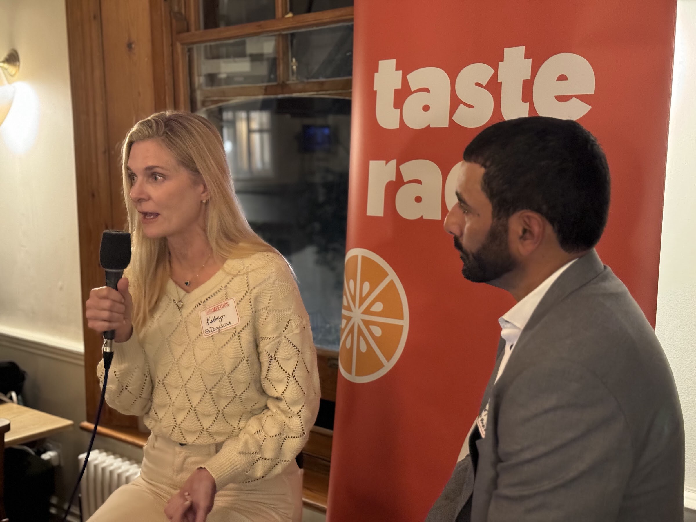 Blonde woman in a cream sweater with a microphone speaking to the interviewer in front of the Taste Radio banner.