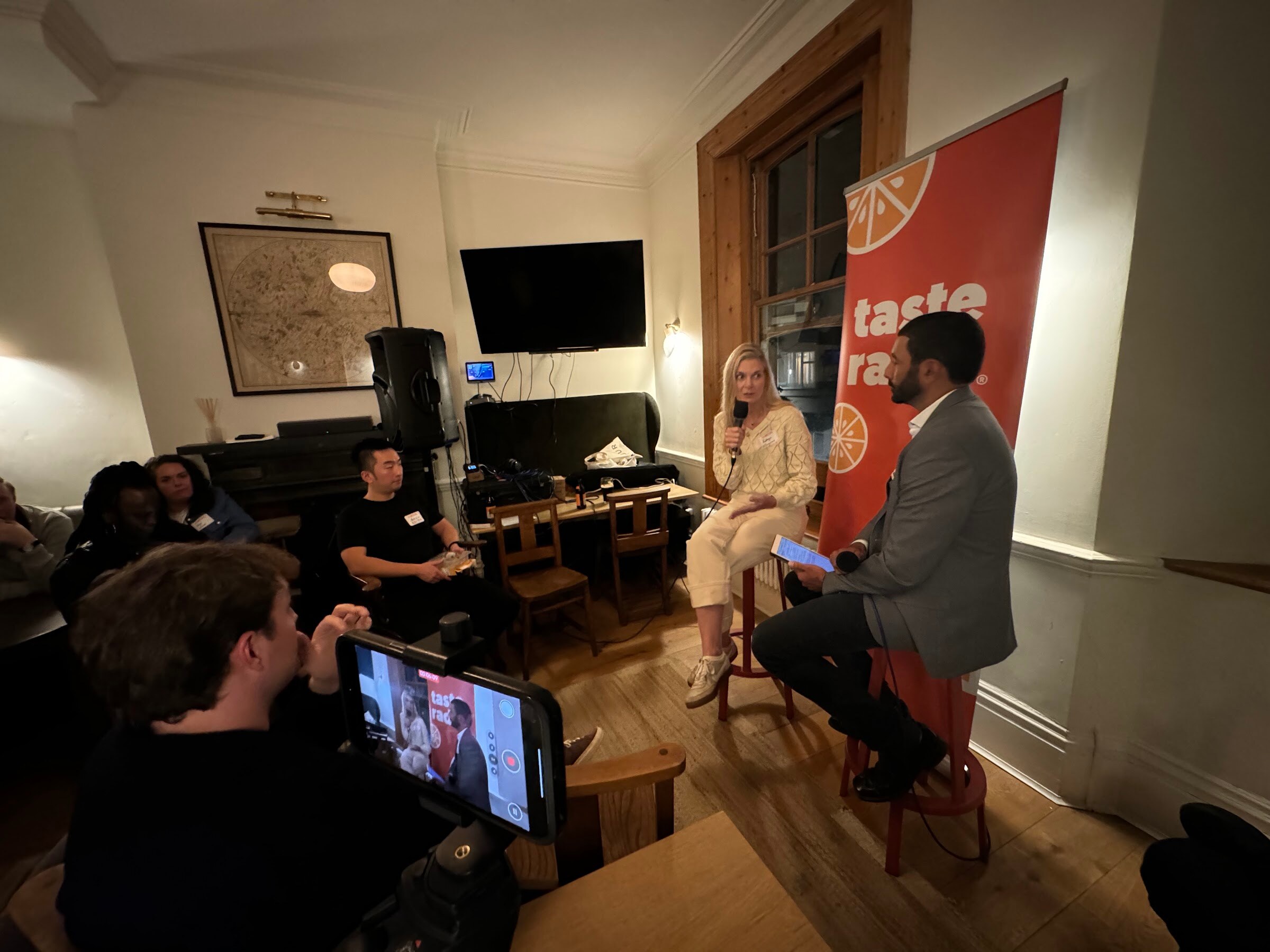 Wide view of the interview corner with a phone on a stand showing the live feed in front, and the interviewer and guest on stools.
