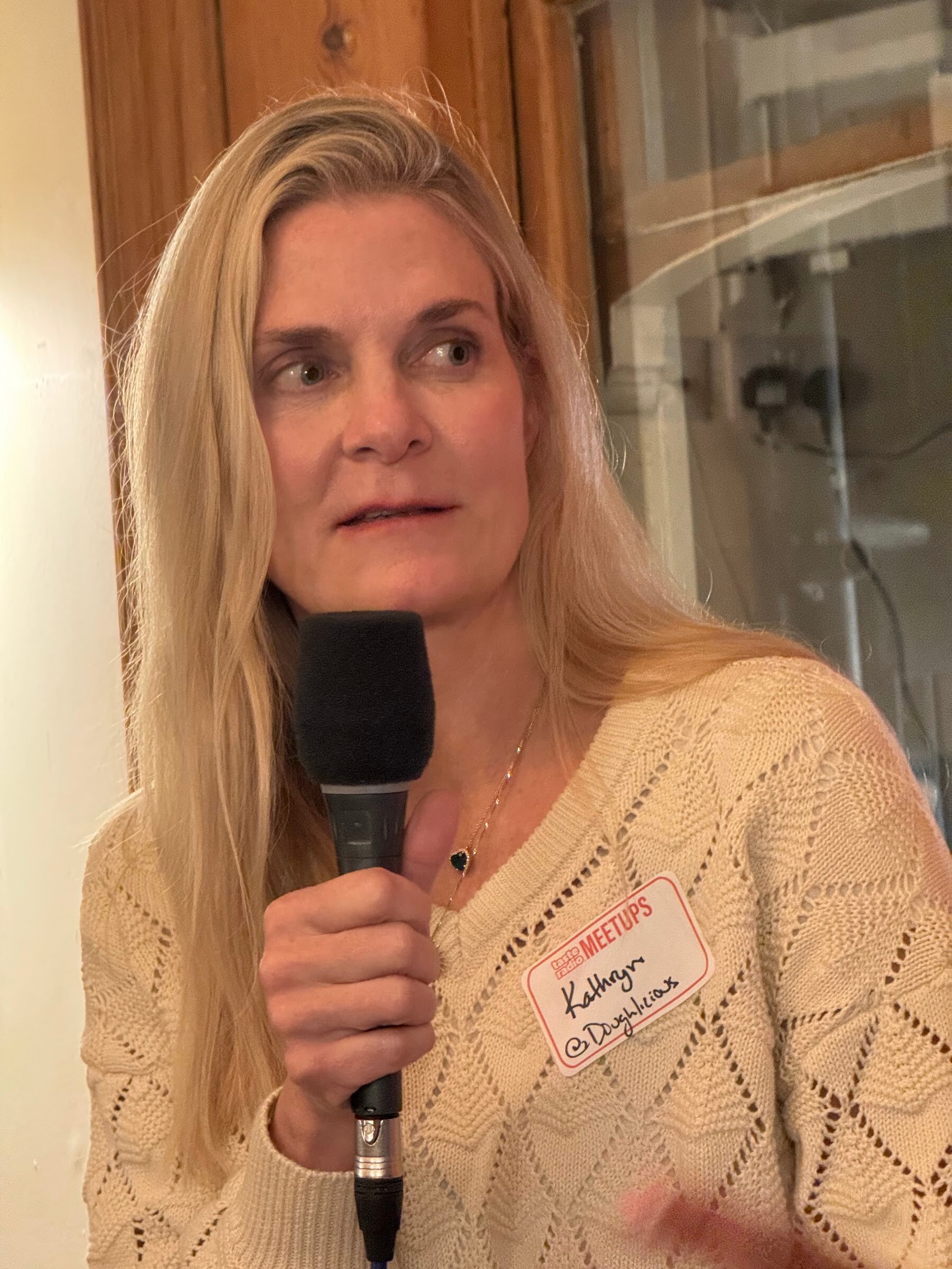 Close-up of a blonde woman in a cream sweater holding a microphone, with a name tag.