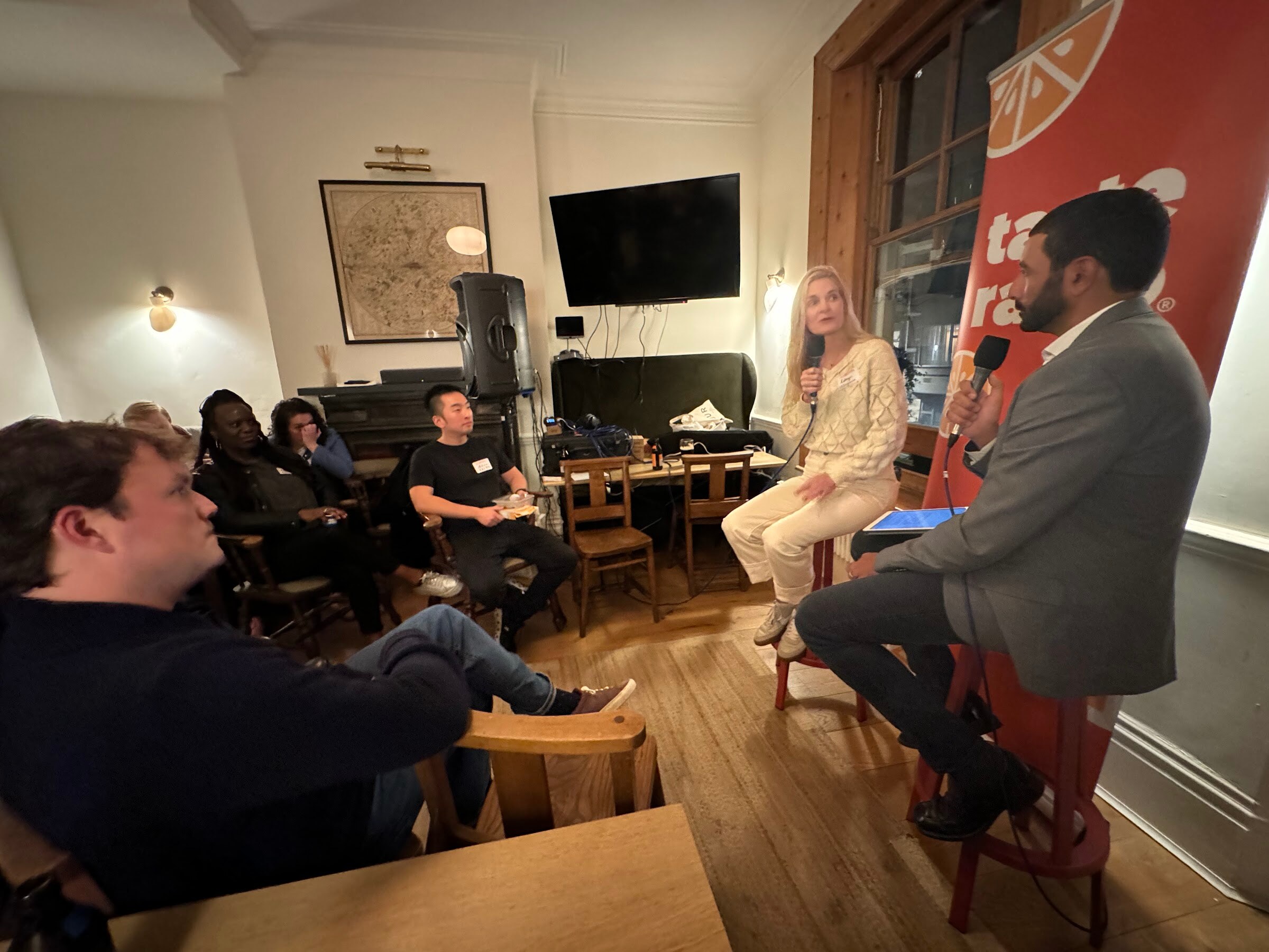 Wide view of the room with seated attendees on the left and the interviewer and guest on stools in front of a Taste Radio banner.
