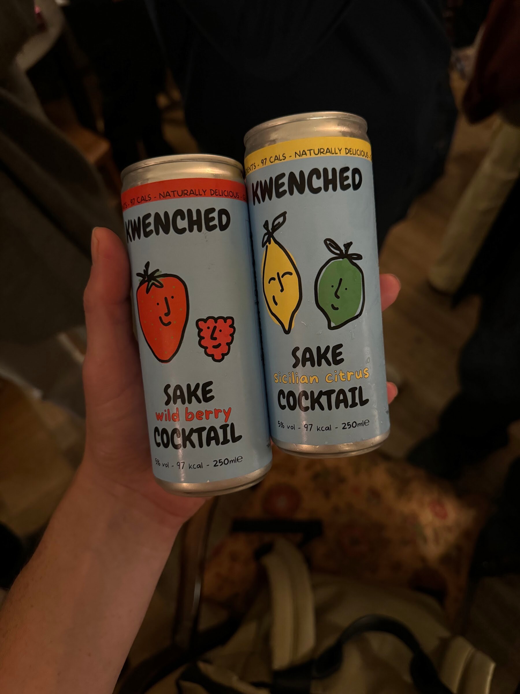 Hand holding two cans of Kwenched sake cocktail, wild berry and Sicilian citrus.