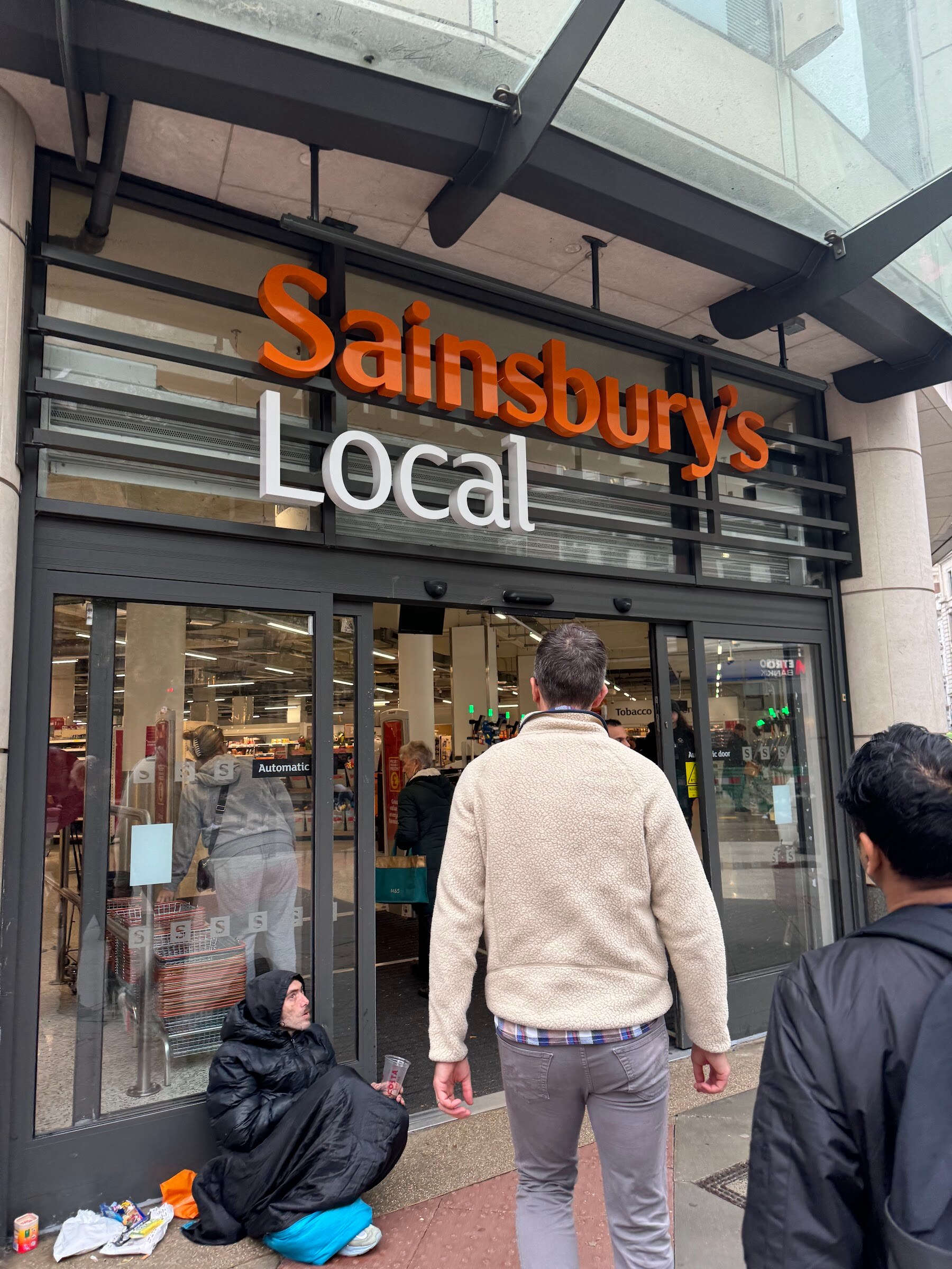 Man in a cream fleece walking into a Sainsbury's Local entrance on a London street.