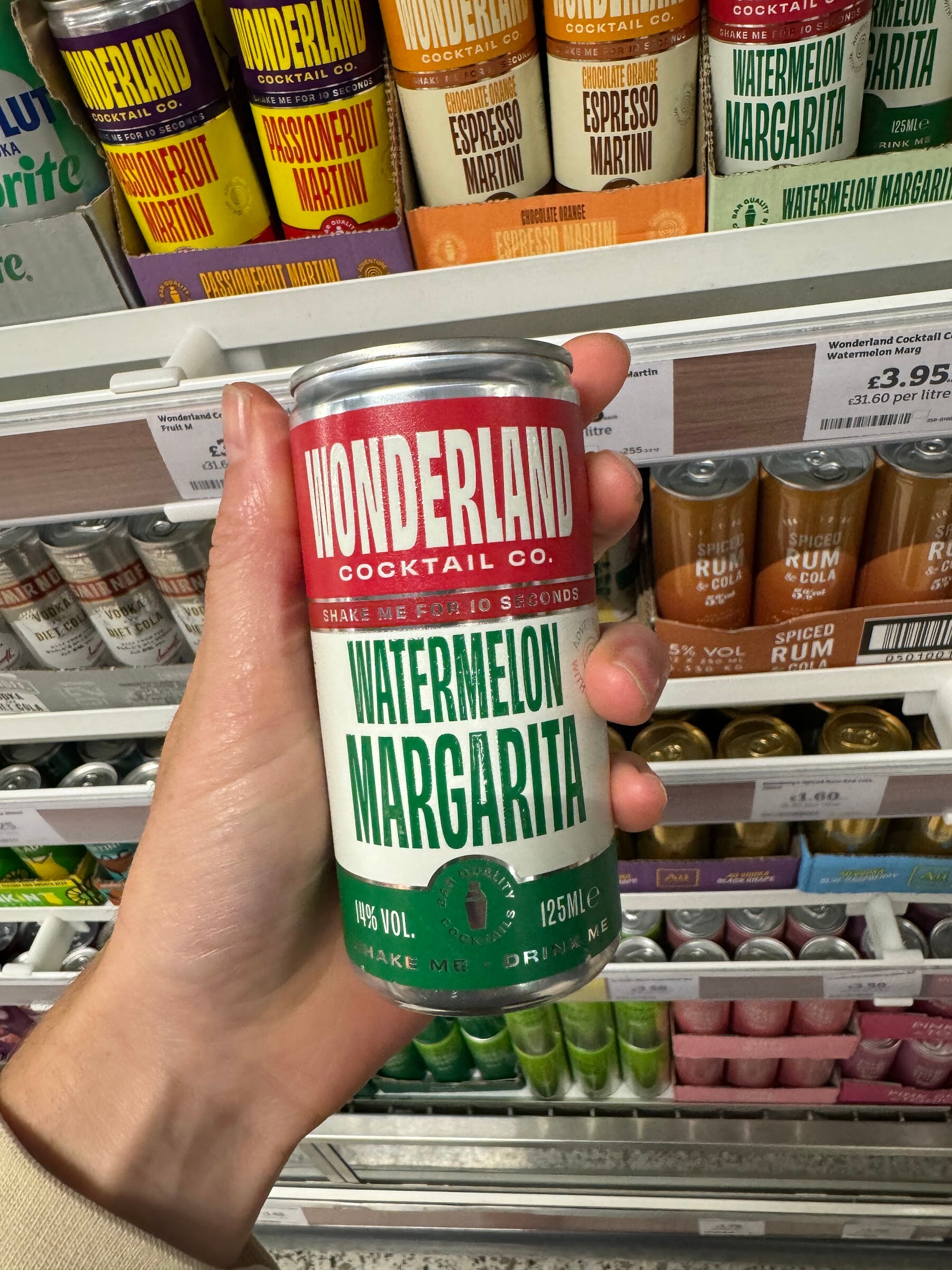 Hand holding a Wonderland Cocktail Co Watermelon Margarita can in front of a shelf of canned cocktails priced in pounds.