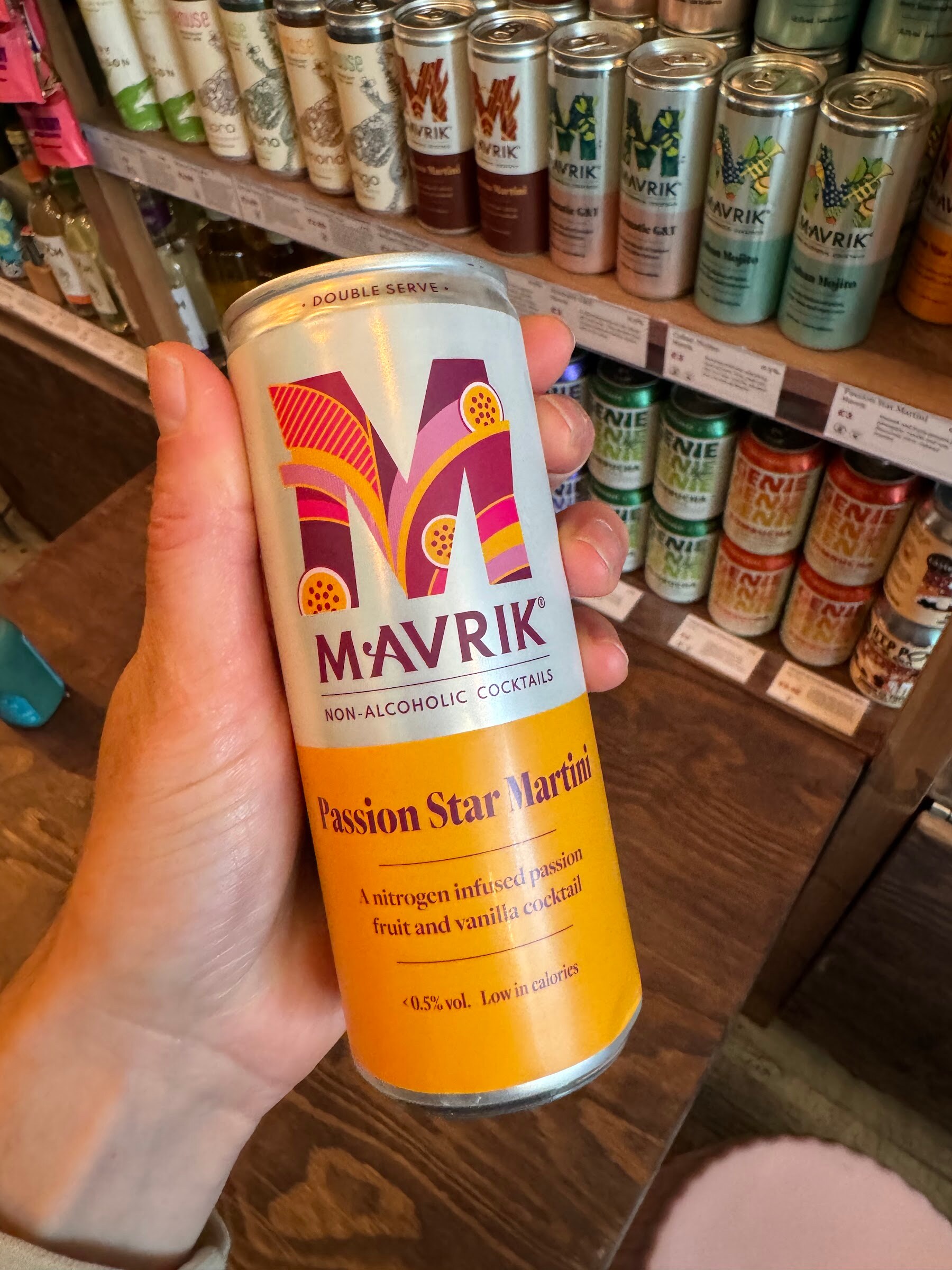 Hand holding a Mavrik non-alcoholic Passion Star Martini can in front of shelves of canned drinks.