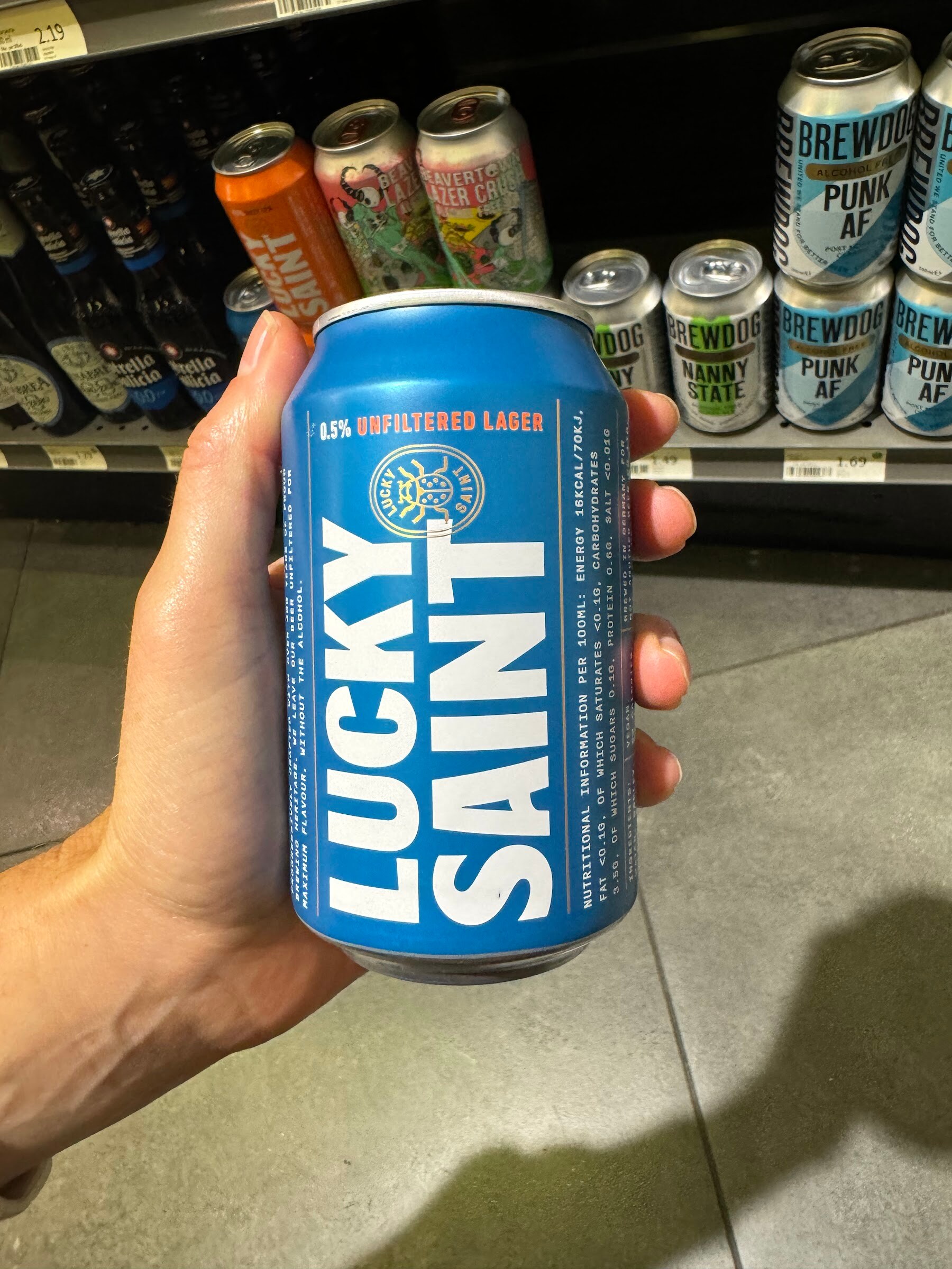 Hand holding a blue Lucky Saint unfiltered lager can in front of shelves of canned craft beers.