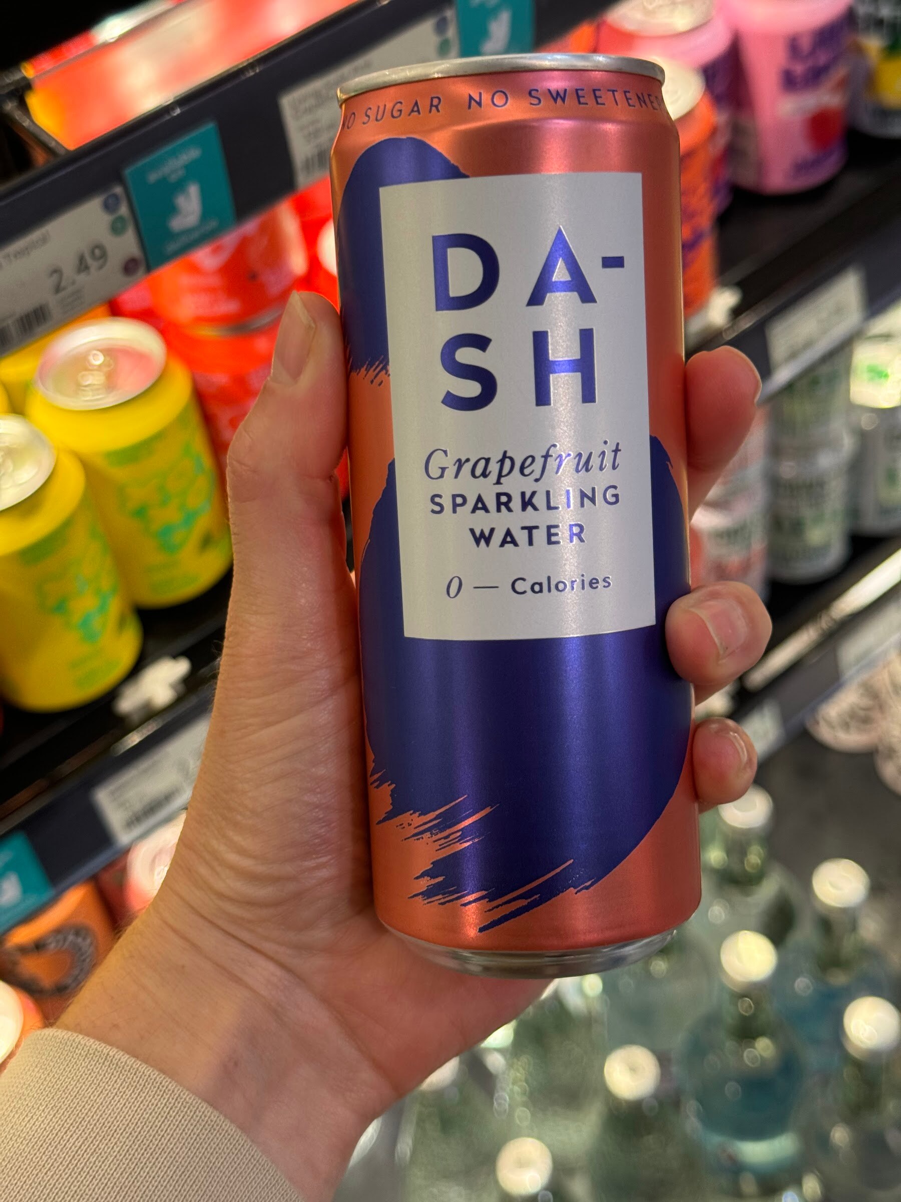 Hand holding a DASH Grapefruit Sparkling Water can in front of a chilled shelf of canned drinks.
