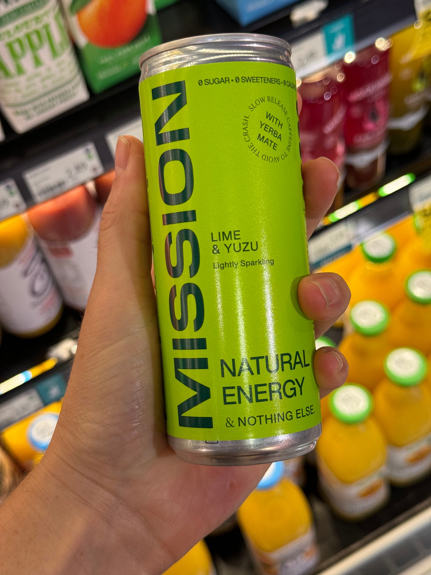 Hand holding a green can of Mission Lime and Yuzu natural energy drink in a chilled aisle.