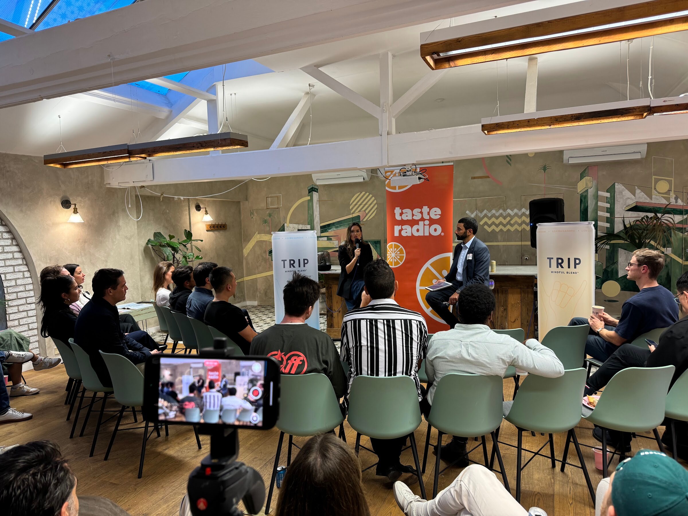 Interviewer and a woman guest on stools in front of Taste Radio and TRIP banners, with seated attendees and a phone on a stand in the foreground.