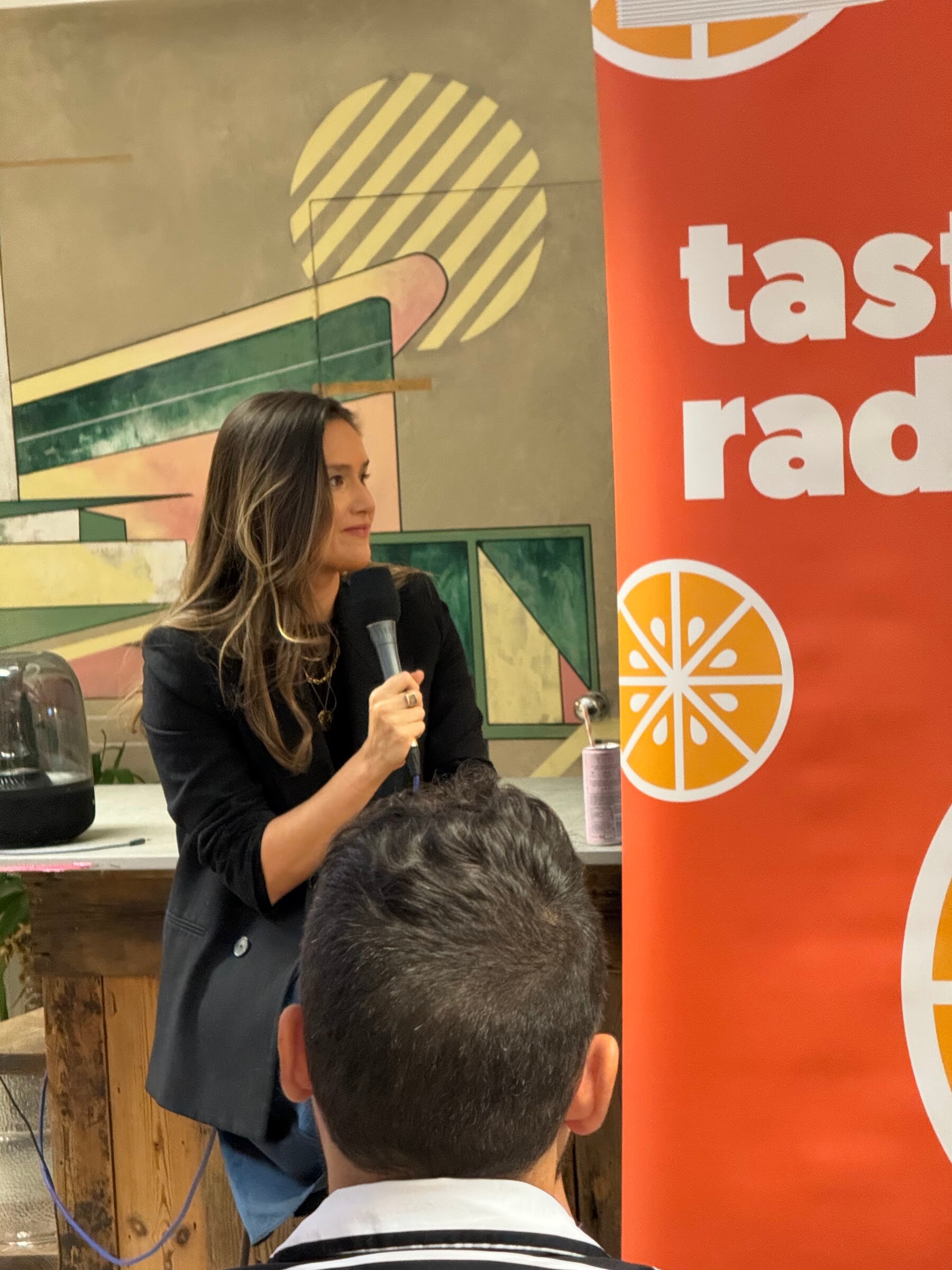 Woman in a black blazer with long hair holding a microphone beside a Taste Radio banner.