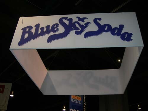 A white hanging rectangular sign with the words Blue Sky Soda in blue script is suspended from the ceiling of the exhibit hall, with an Expo East banner visible below.
