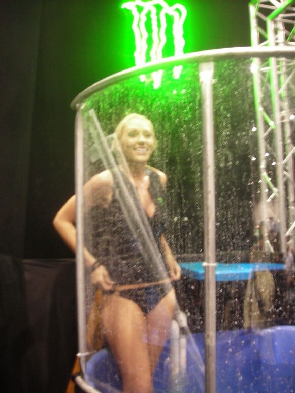 A smiling blonde woman in a dark swimsuit-style top stands inside a clear dunk tank with water falling around her, beneath a glowing green Monster logo.