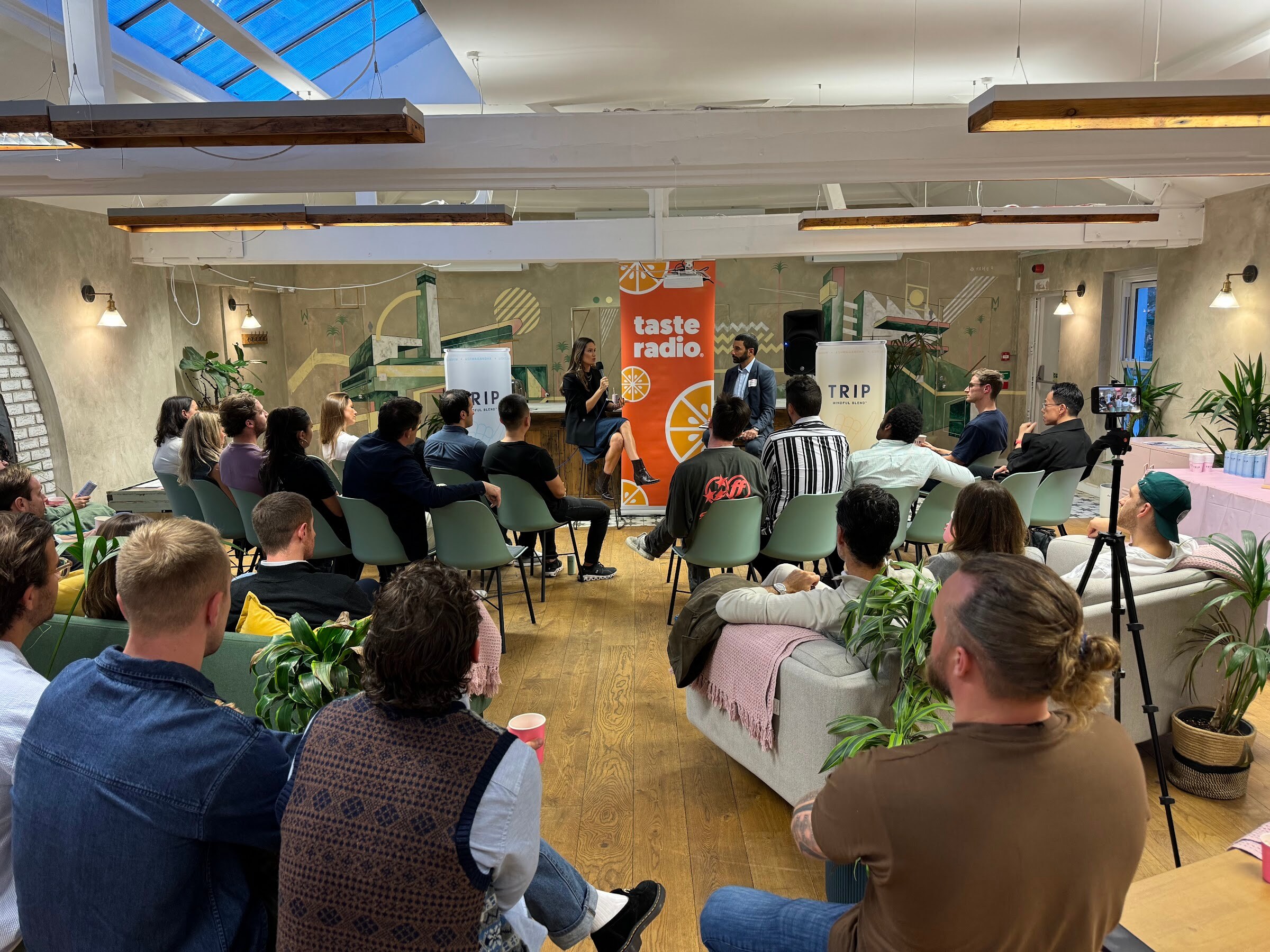 Wide view from the back of the room with attendees on sofas and chairs facing the Taste Radio banner under a skylight.