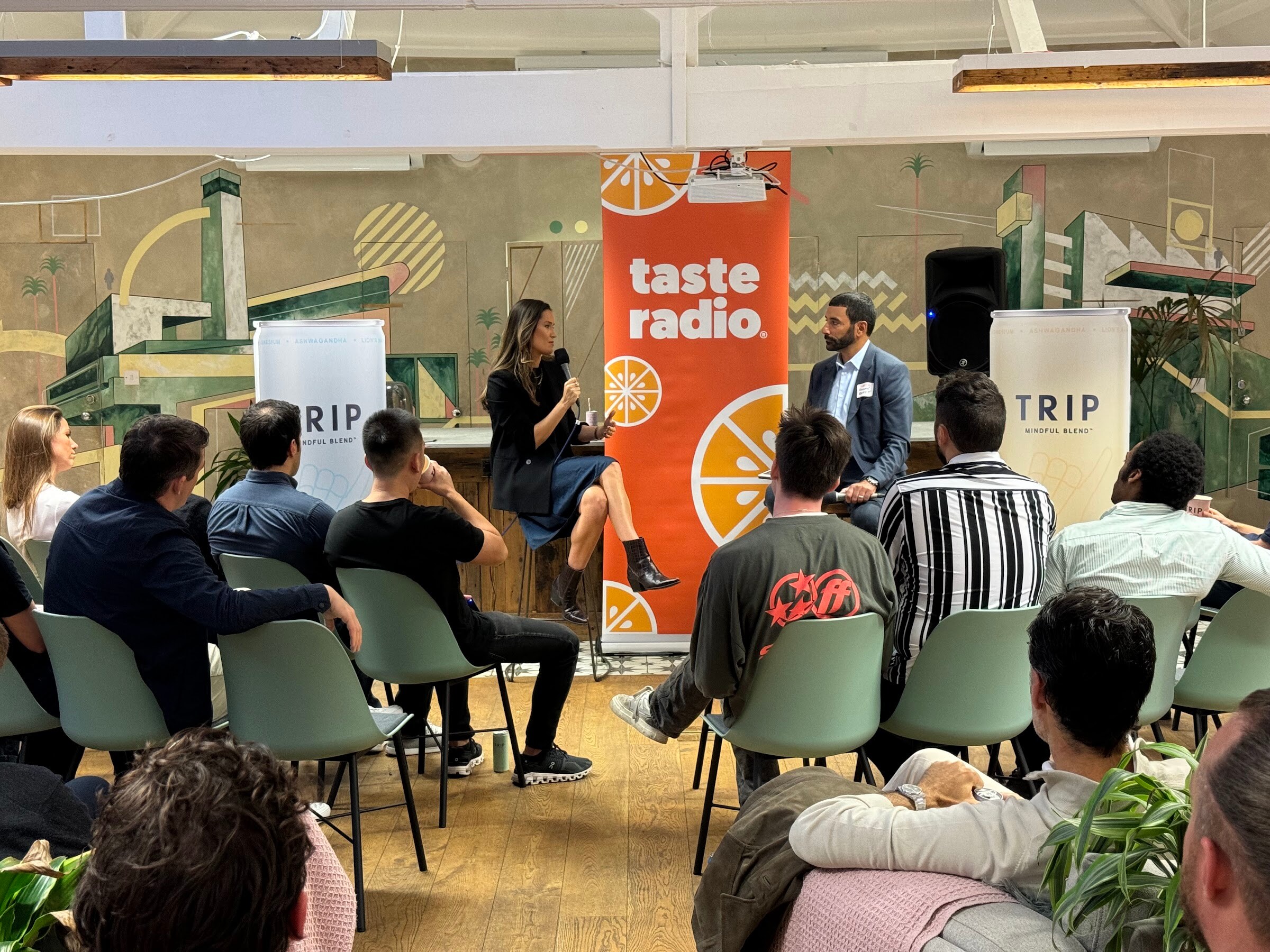 Woman in a black blazer and the Taste Radio interviewer seated between Taste Radio and TRIP banners with attendees in front.