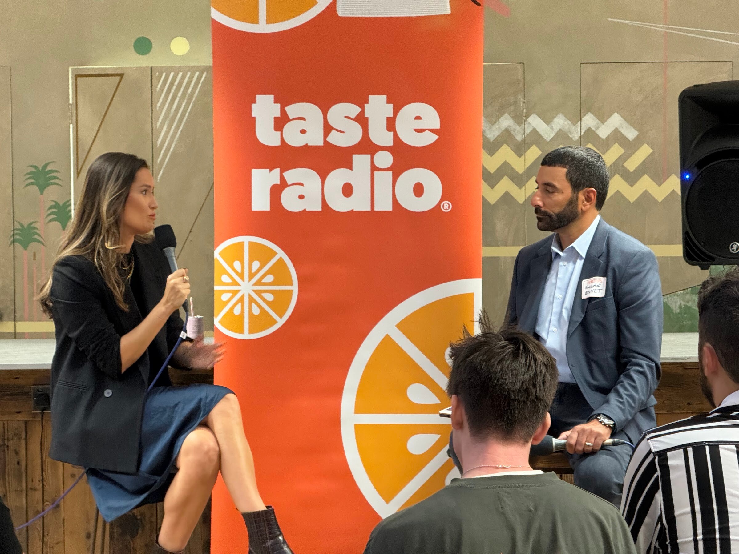 Woman in a black blazer holding a microphone speaking with the interviewer in a blue blazer beside a Taste Radio banner.