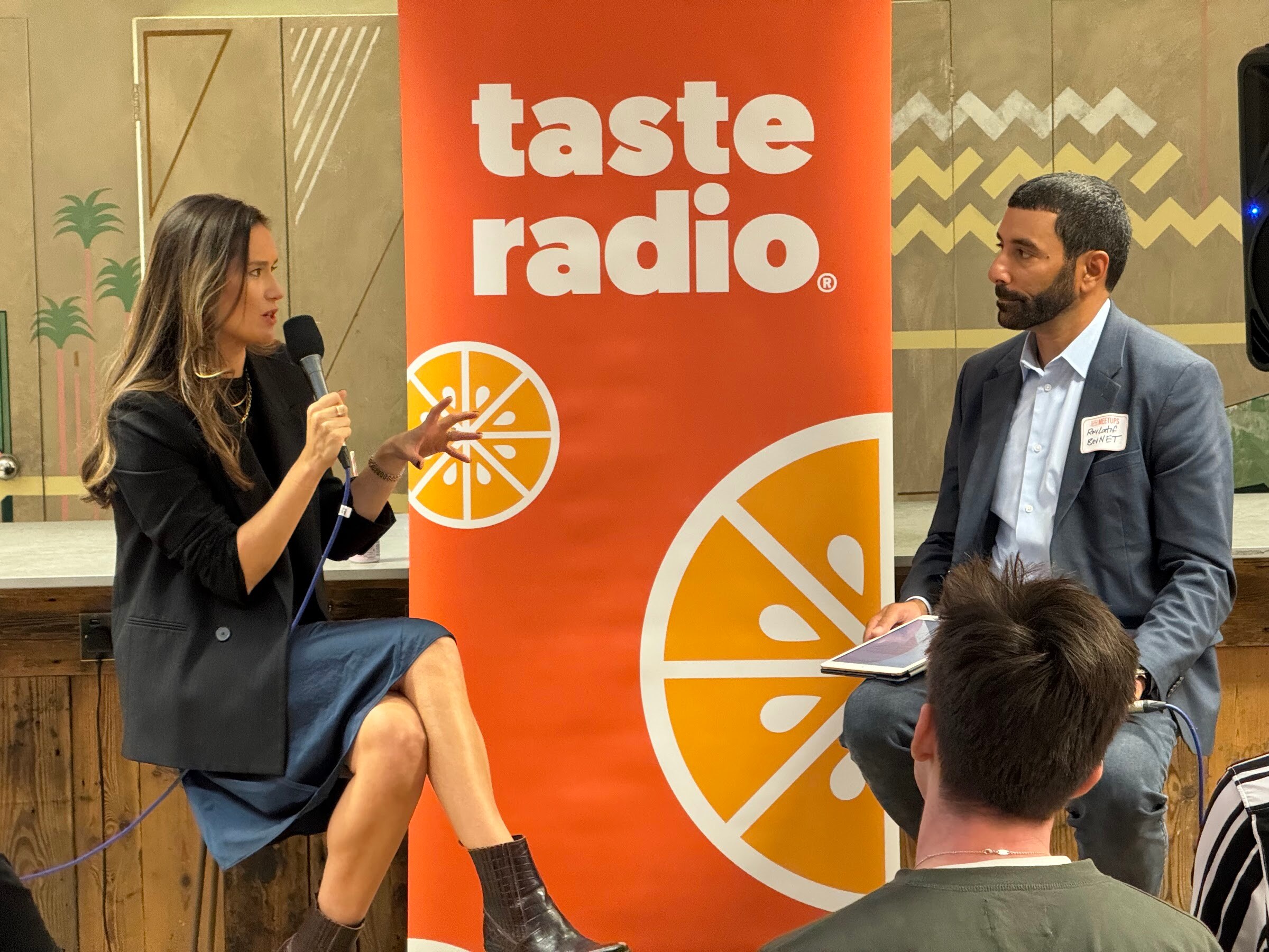 Woman in a black blazer gesturing as she speaks, with the interviewer in a blue blazer holding a tablet next to the Taste Radio banner.