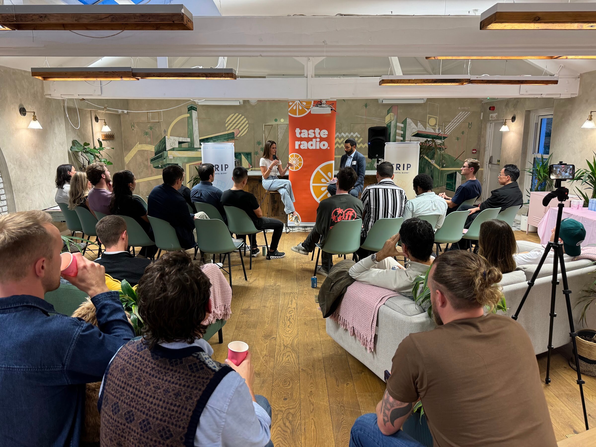 Wide view of the TRIP HQ room with many seated attendees watching the interviewer and guest in front of a Taste Radio banner.