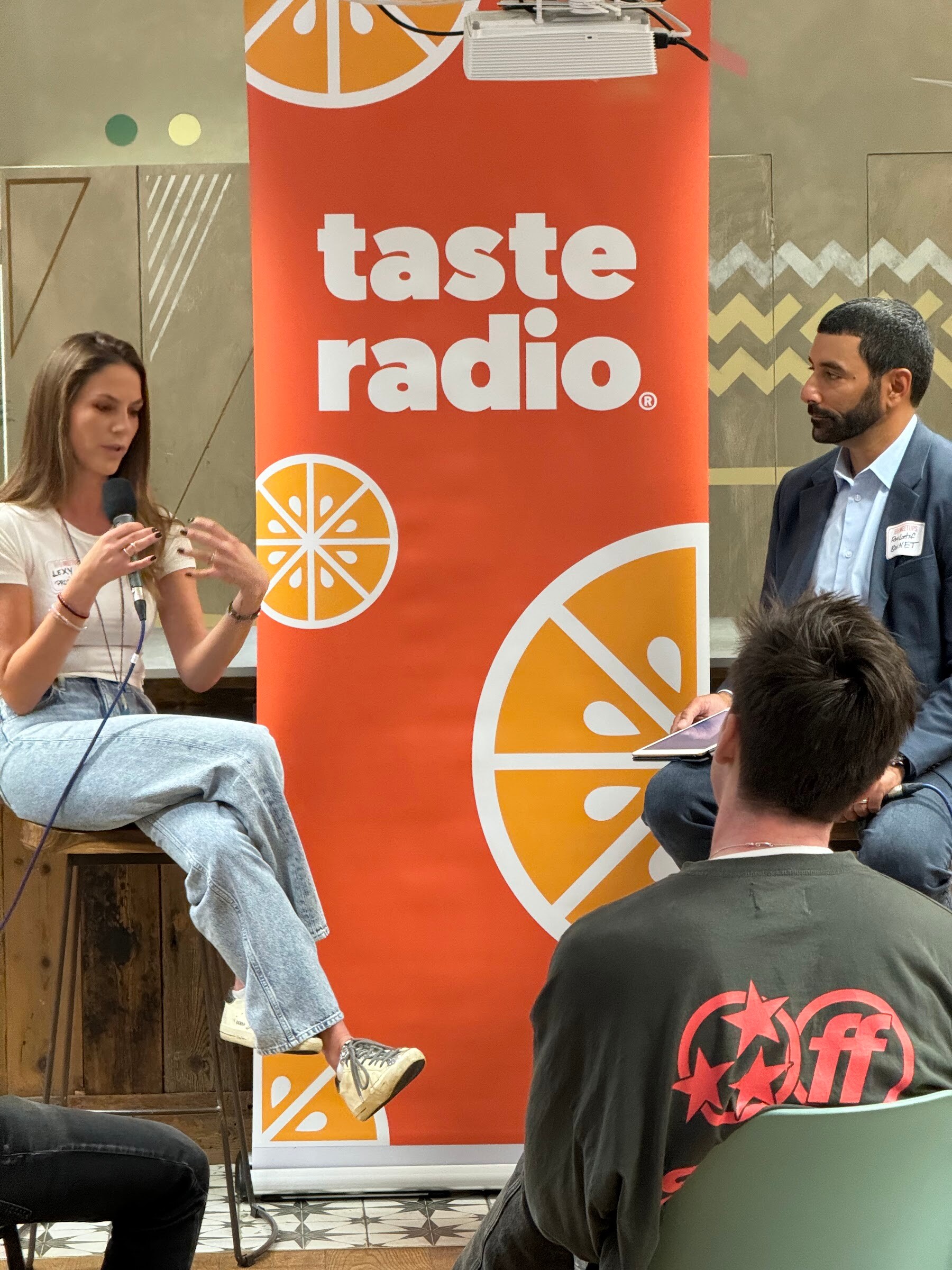 Woman in a white tee and light jeans holding a microphone on a stool beside a Taste Radio banner.