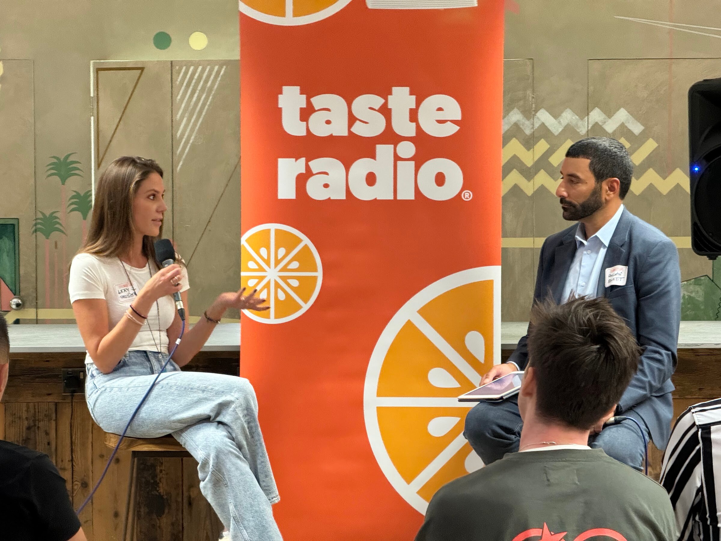 Woman in a white tee and light jeans with a microphone, speaking to the interviewer in a blue blazer in front of the Taste Radio banner.