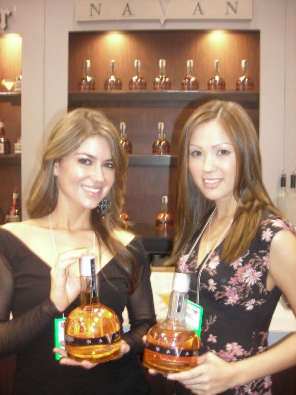 Two smiling women each hold a rounded bottle of amber Navan liqueur in front of a shelf displaying more Navan bottles under a Navan sign.