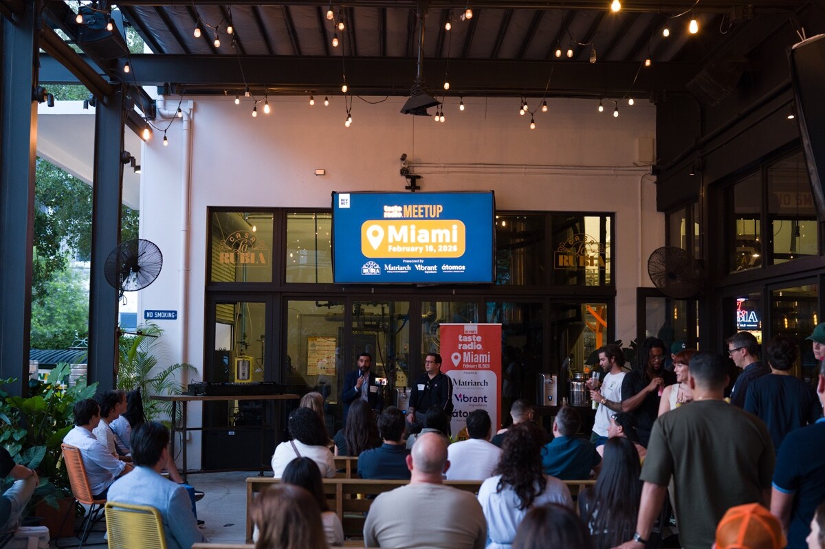 Wide view of a crowd on a covered patio facing a stage with a screen reading Taste Radio Meetup Miami.
