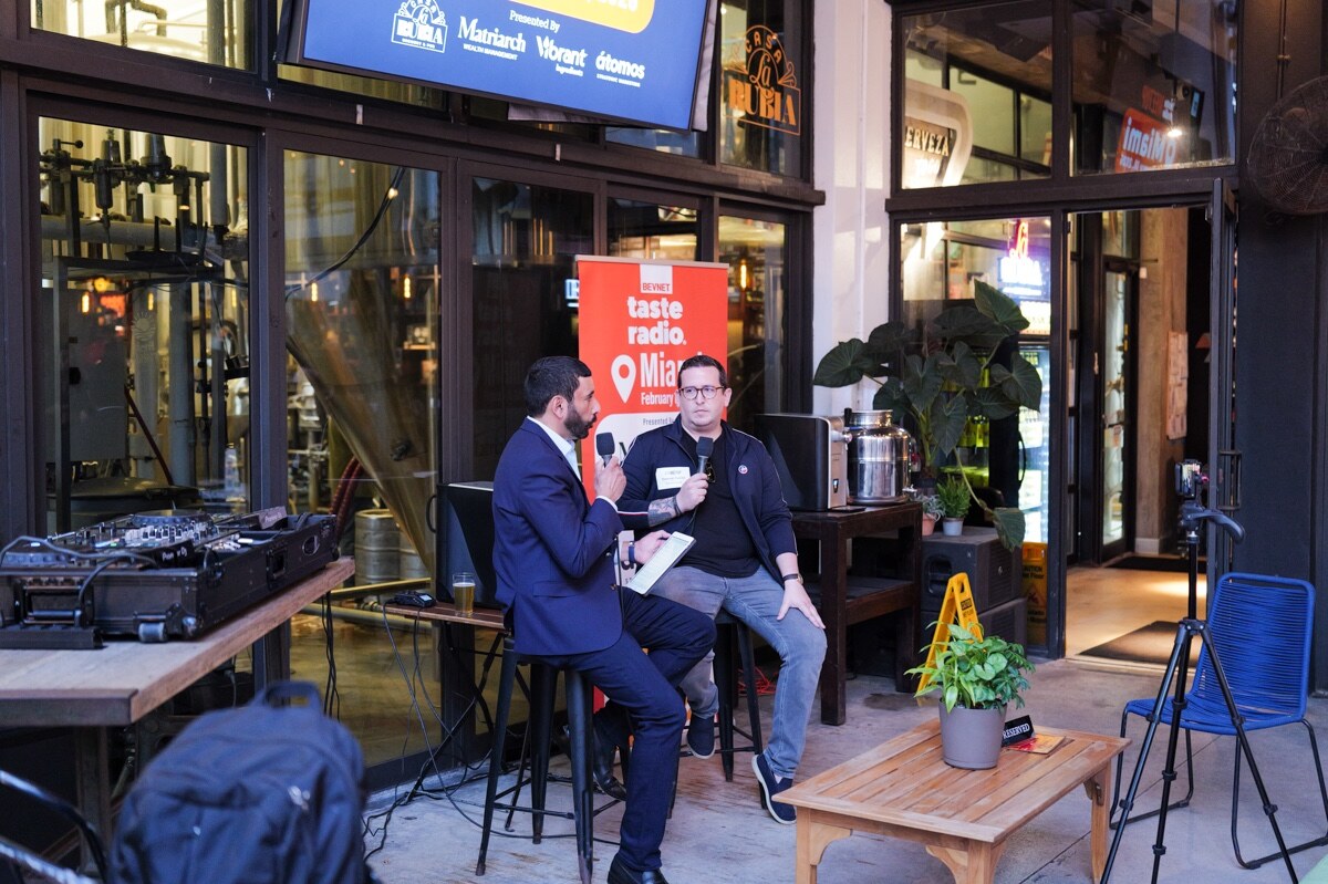 Host in a navy suit and a guest in black talk into microphones on stools next to the Taste Radio Miami banner.