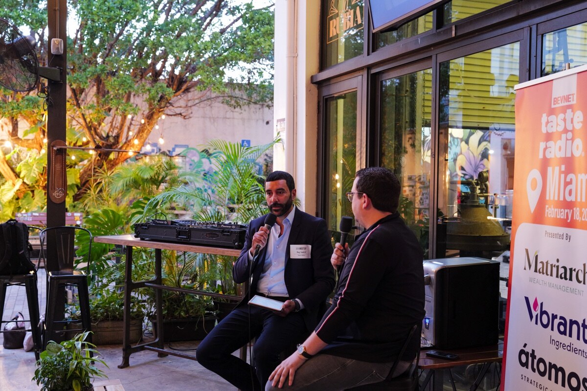 Host and guest on stools with microphones next to the Taste Radio Miami banner, tropical plants behind.