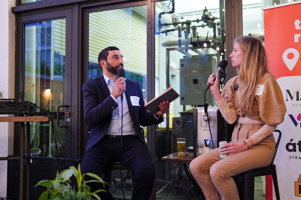 Host in a suit holding a tablet and microphone faces a woman in a tan outfit with a microphone.