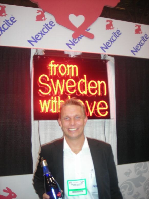 A smiling man in a dark jacket and white shirt wears a badge and holds a blue bottle in front of a red neon sign reading from Sweden with love and Nexcite banners.