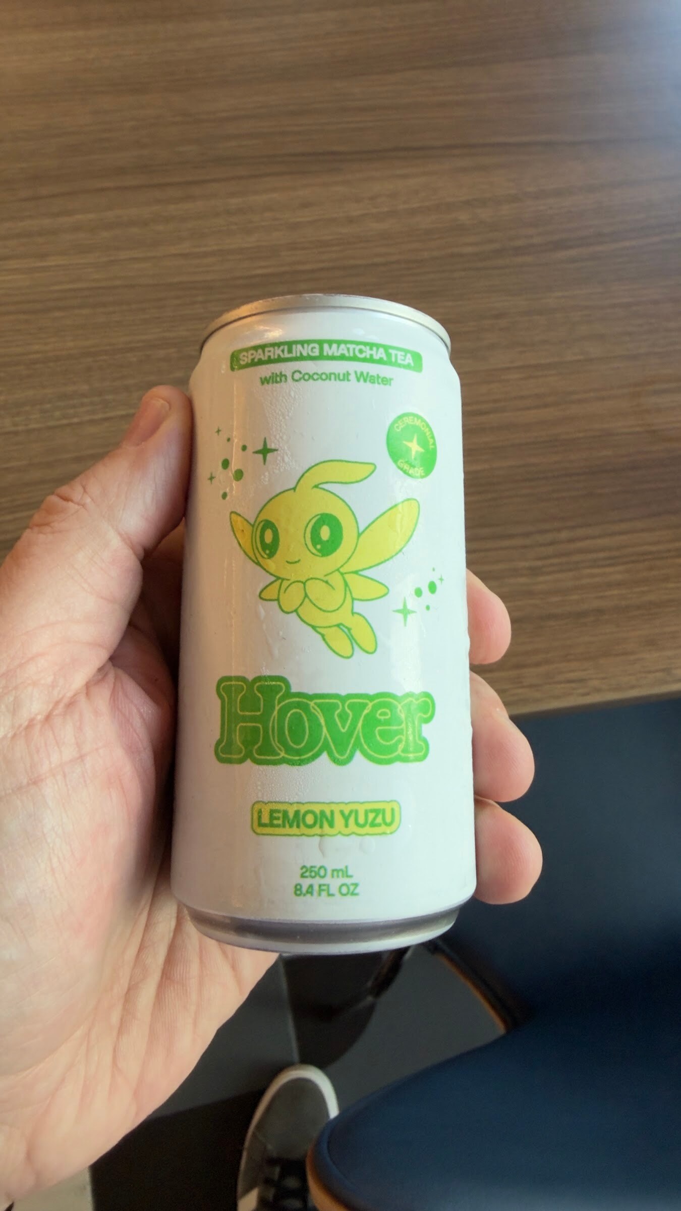 Hand holding a white can of Hover Lemon Yuzu sparkling matcha tea over a wood table.