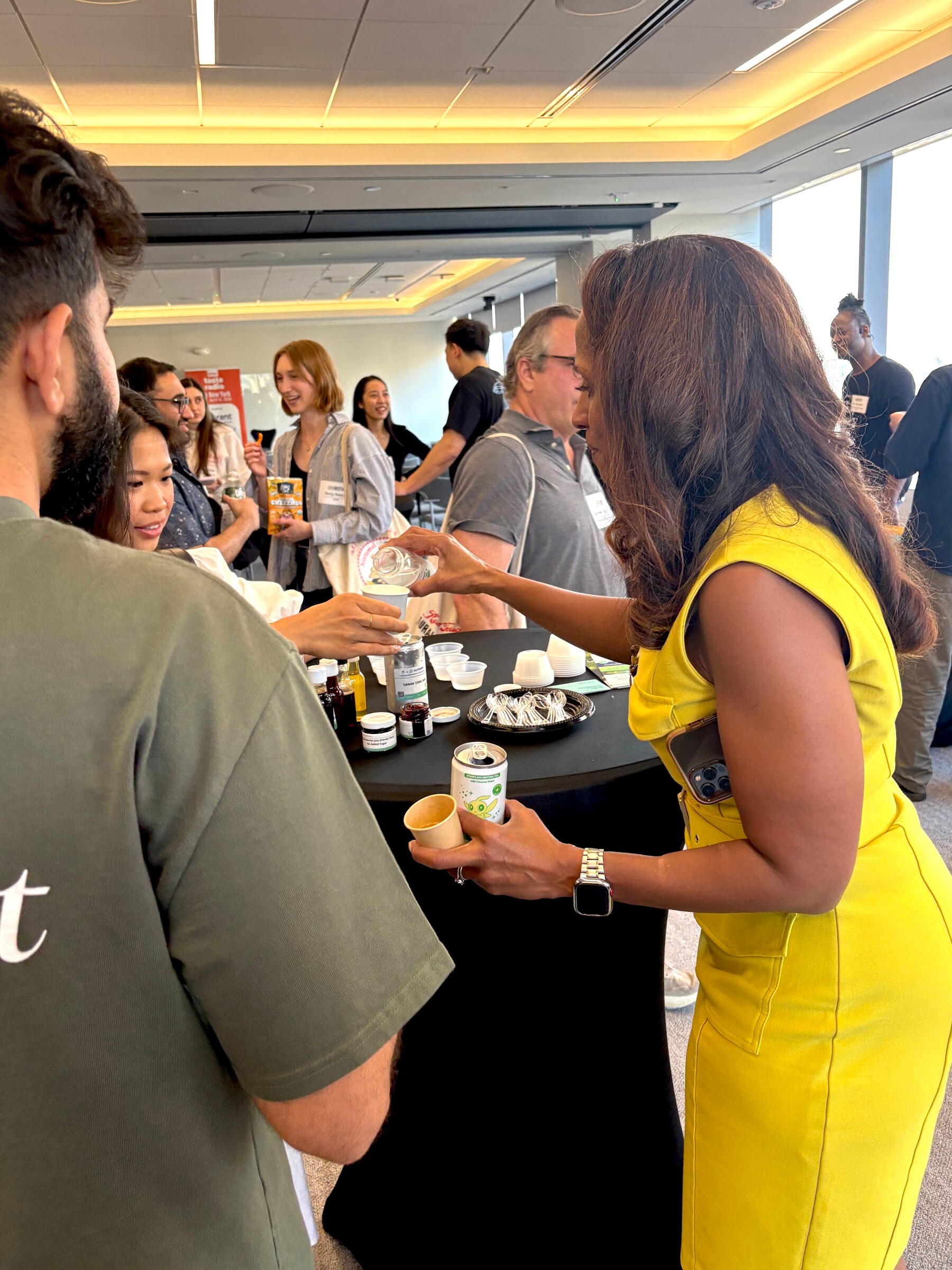 Attendees gather around a black high-top table pouring samples, with more guests behind in a bright office.