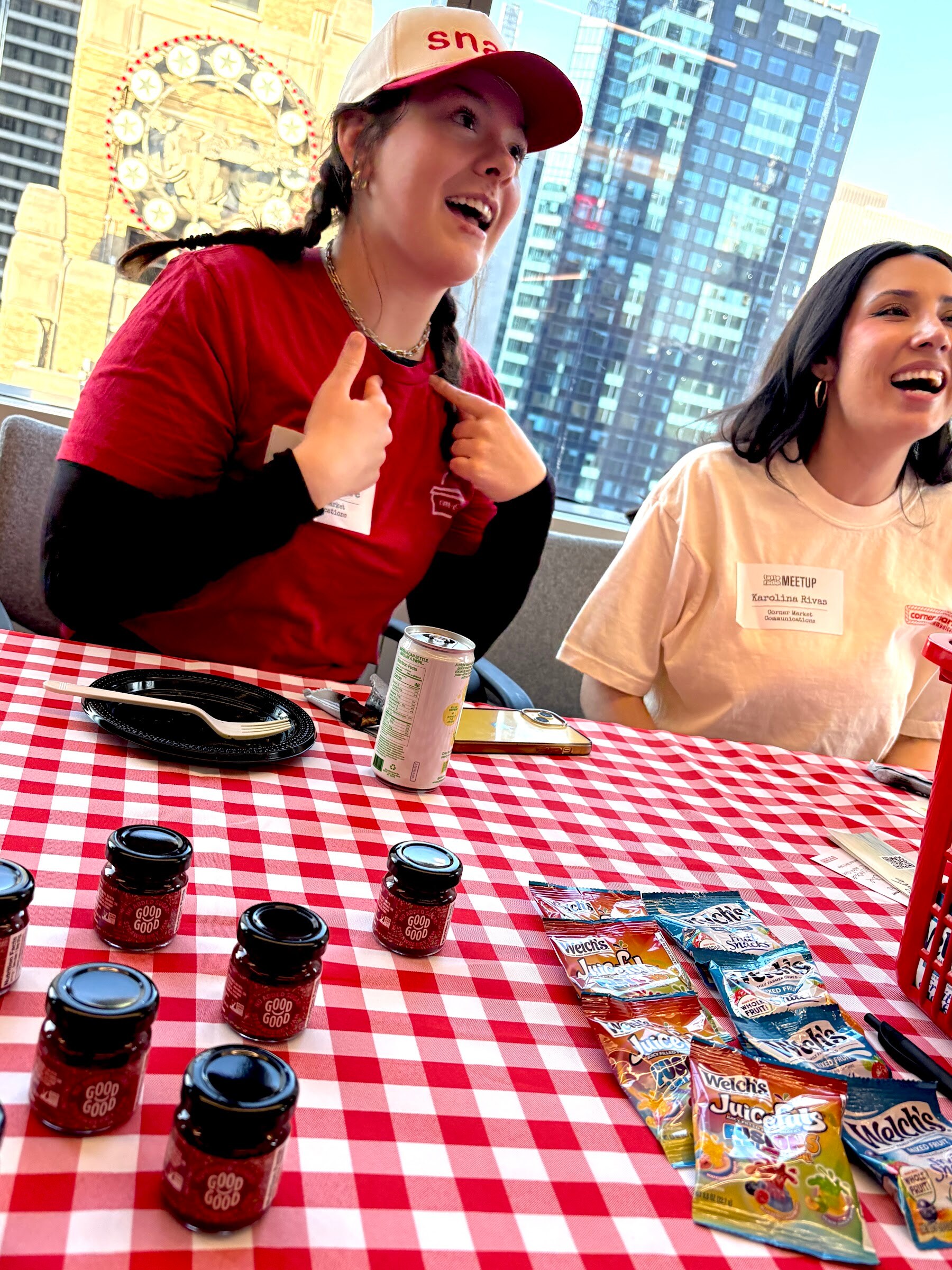 Two women laugh at a red gingham table set with small jam jars and packets of fruit snacks, with skyscrapers through the window.