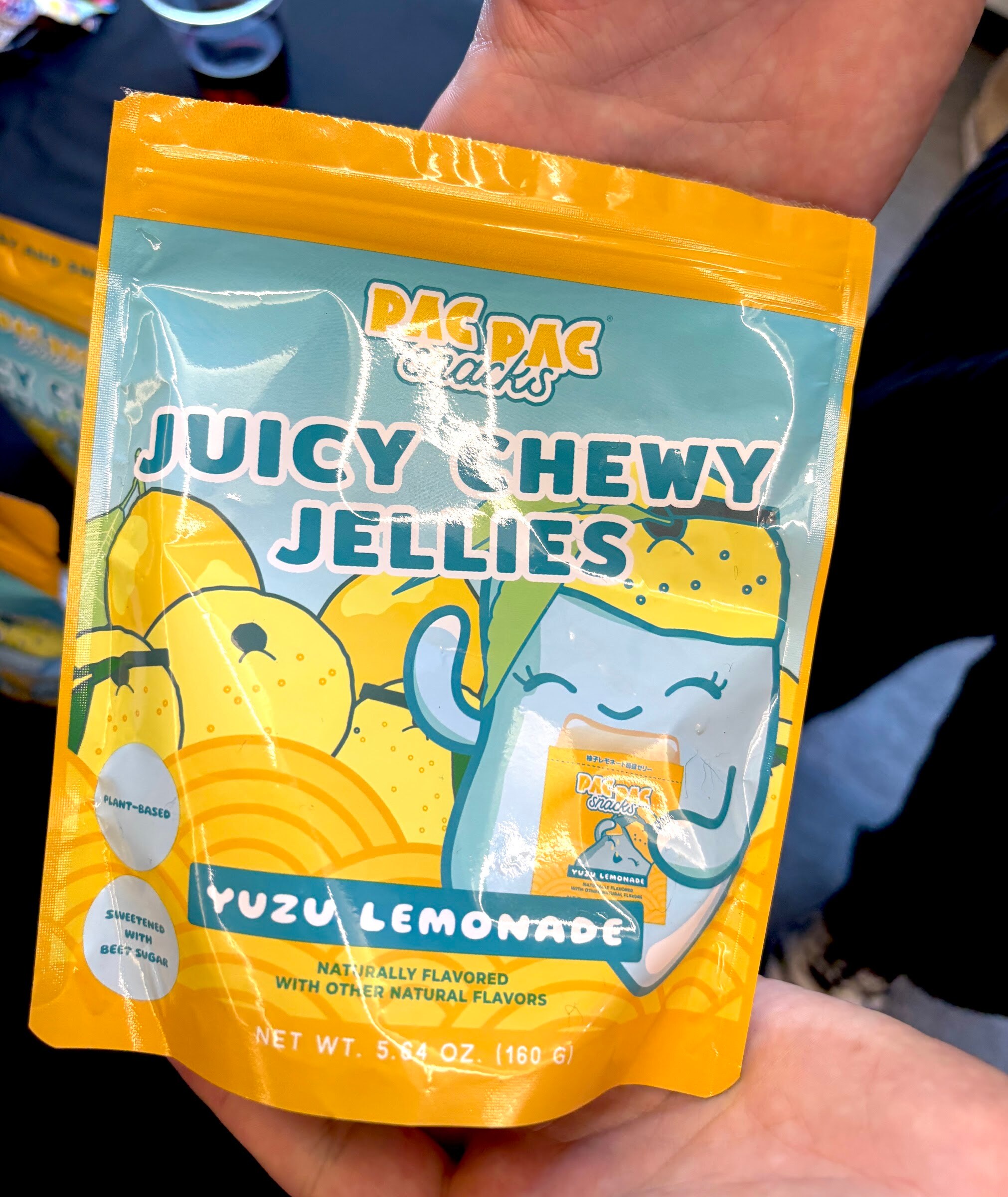 Hand holding a yellow and blue pouch of Pac Pac Snacks Juicy Chewy Jellies, Yuzu Lemonade.