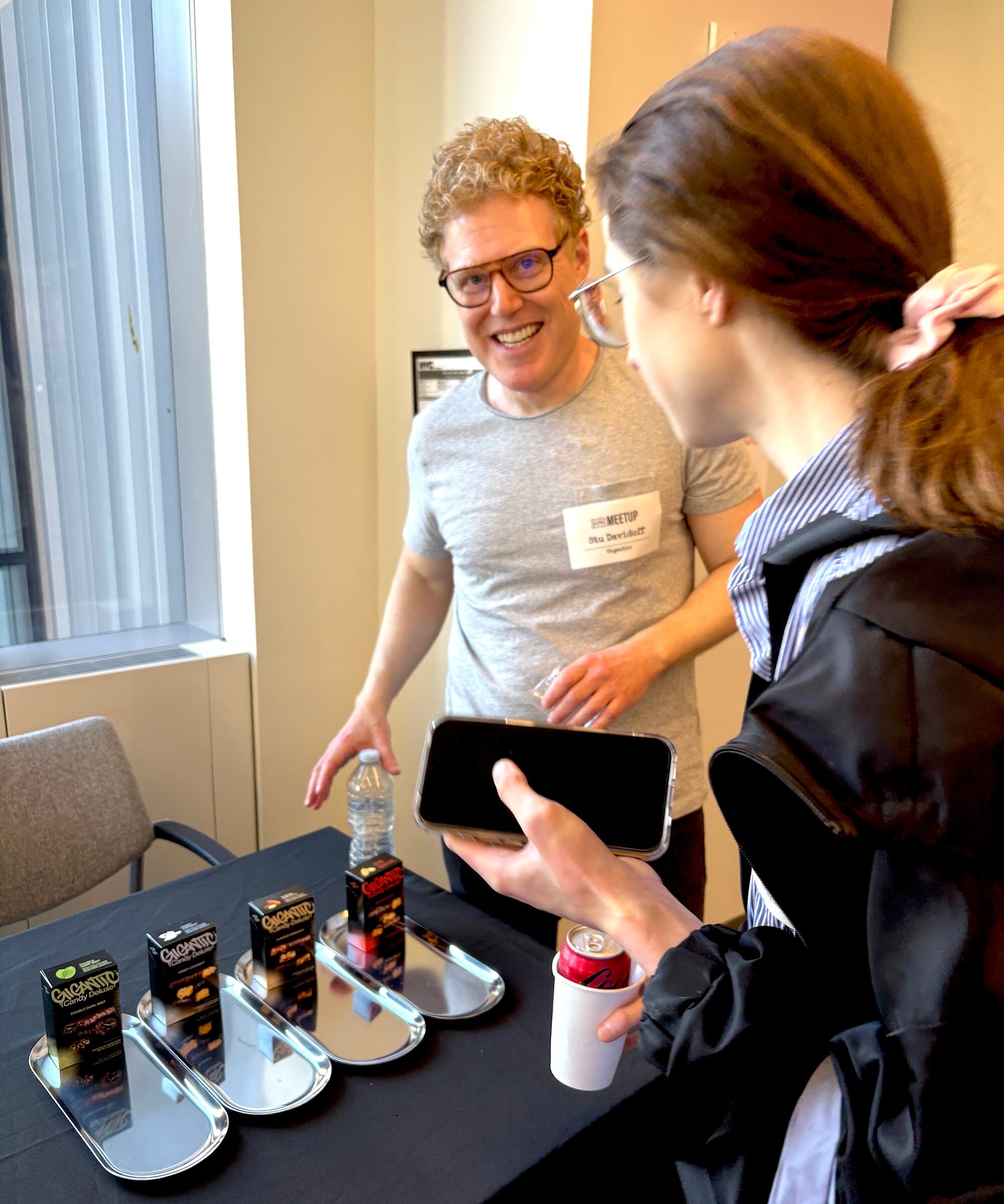 Smiling man in glasses stands behind a table of Gigantic candy boxes on silver trays as an attendee holds up a phone.