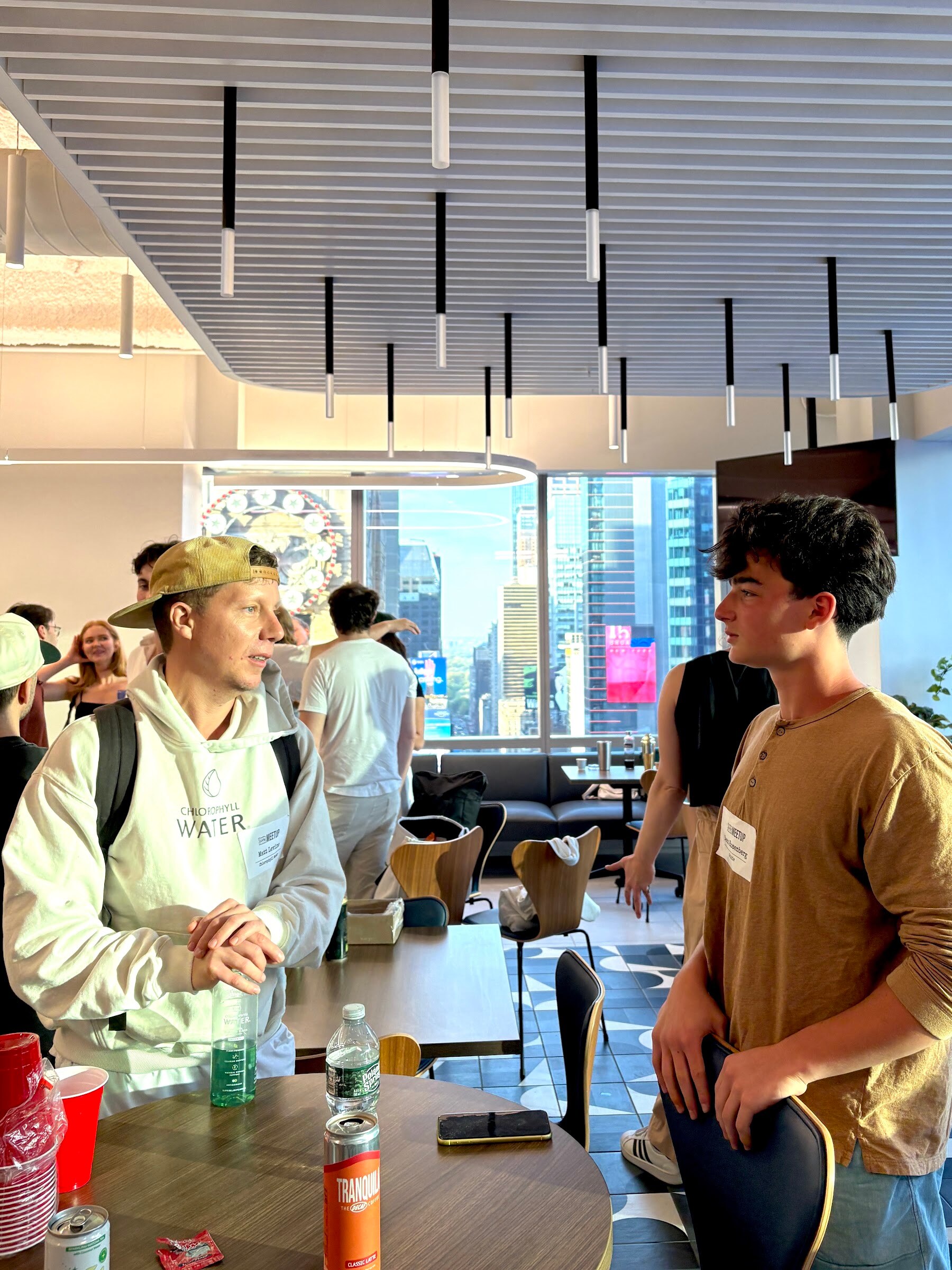 Man in a tan cap and Chlorophyll Water hoodie talks with a young man in a brown henley, with Times Square buildings through the windows.