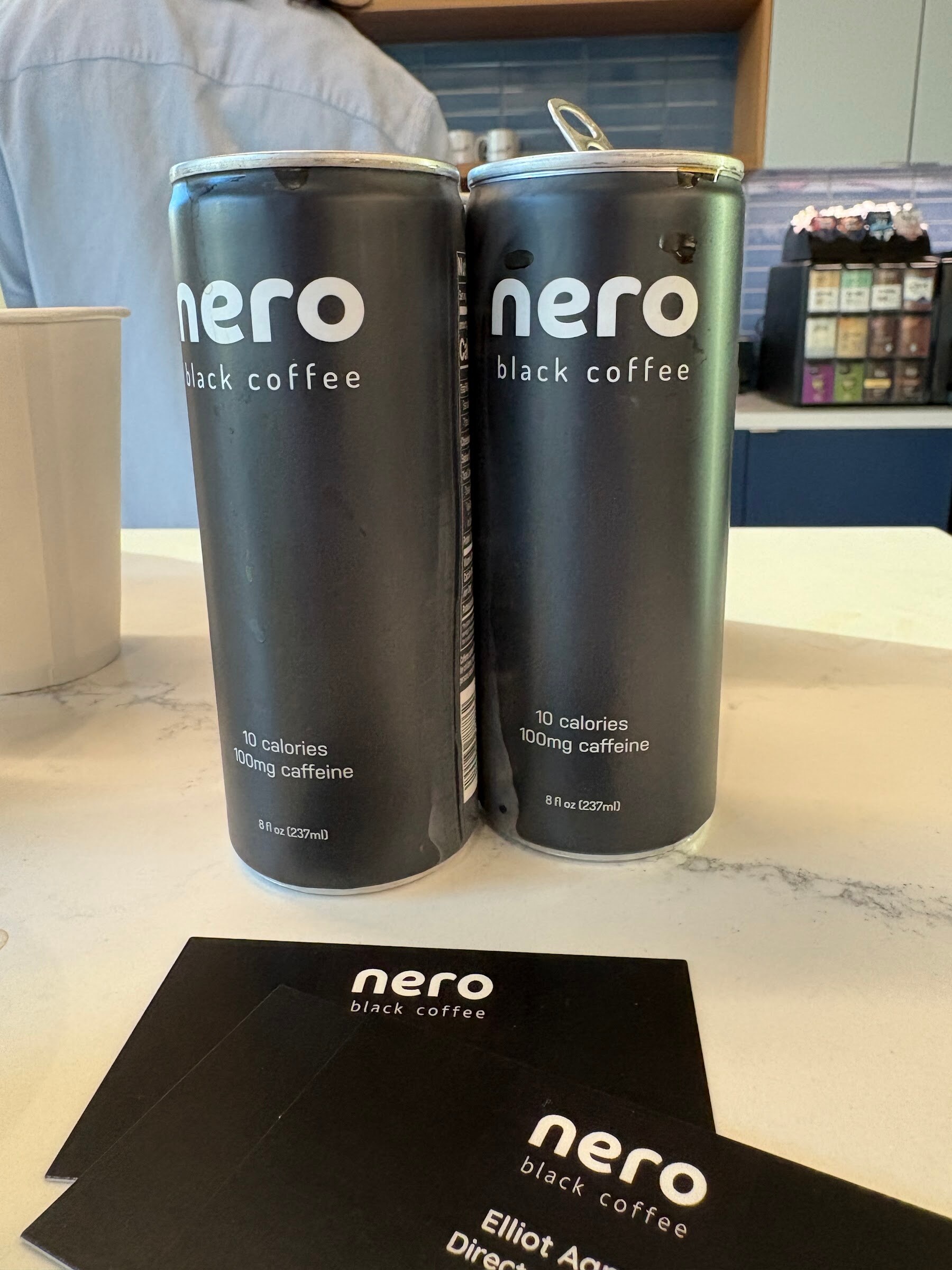Two black cans of Nero black coffee on a white counter beside Nero business cards.
