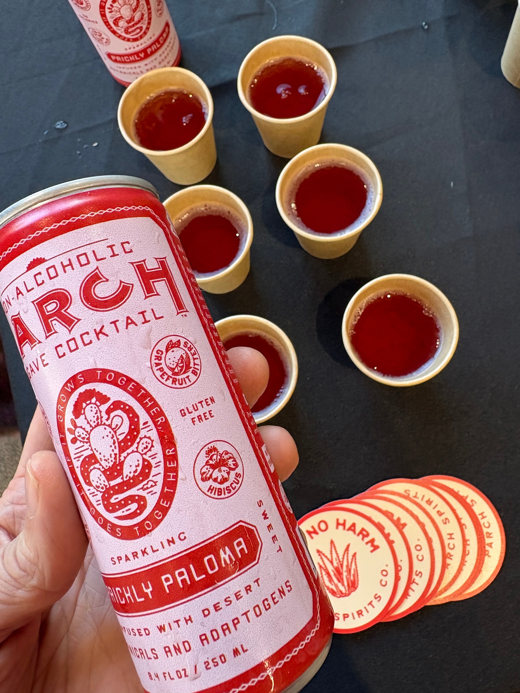 Hand holding a red and white can of Parch Prickly Paloma above sample cups of red drink.