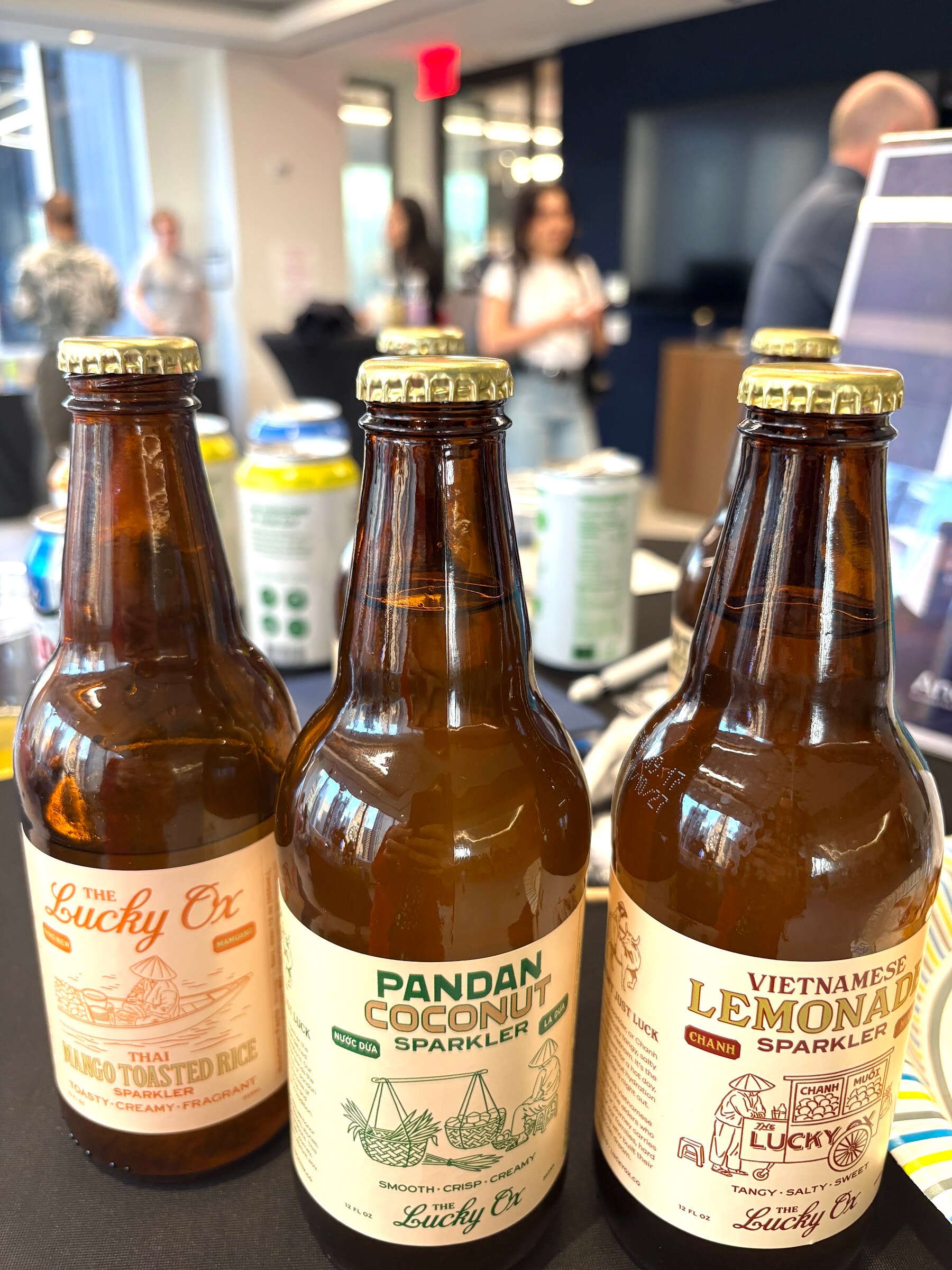 Three amber bottles of The Lucky Ox sparklers on a table with attendees blurred behind.