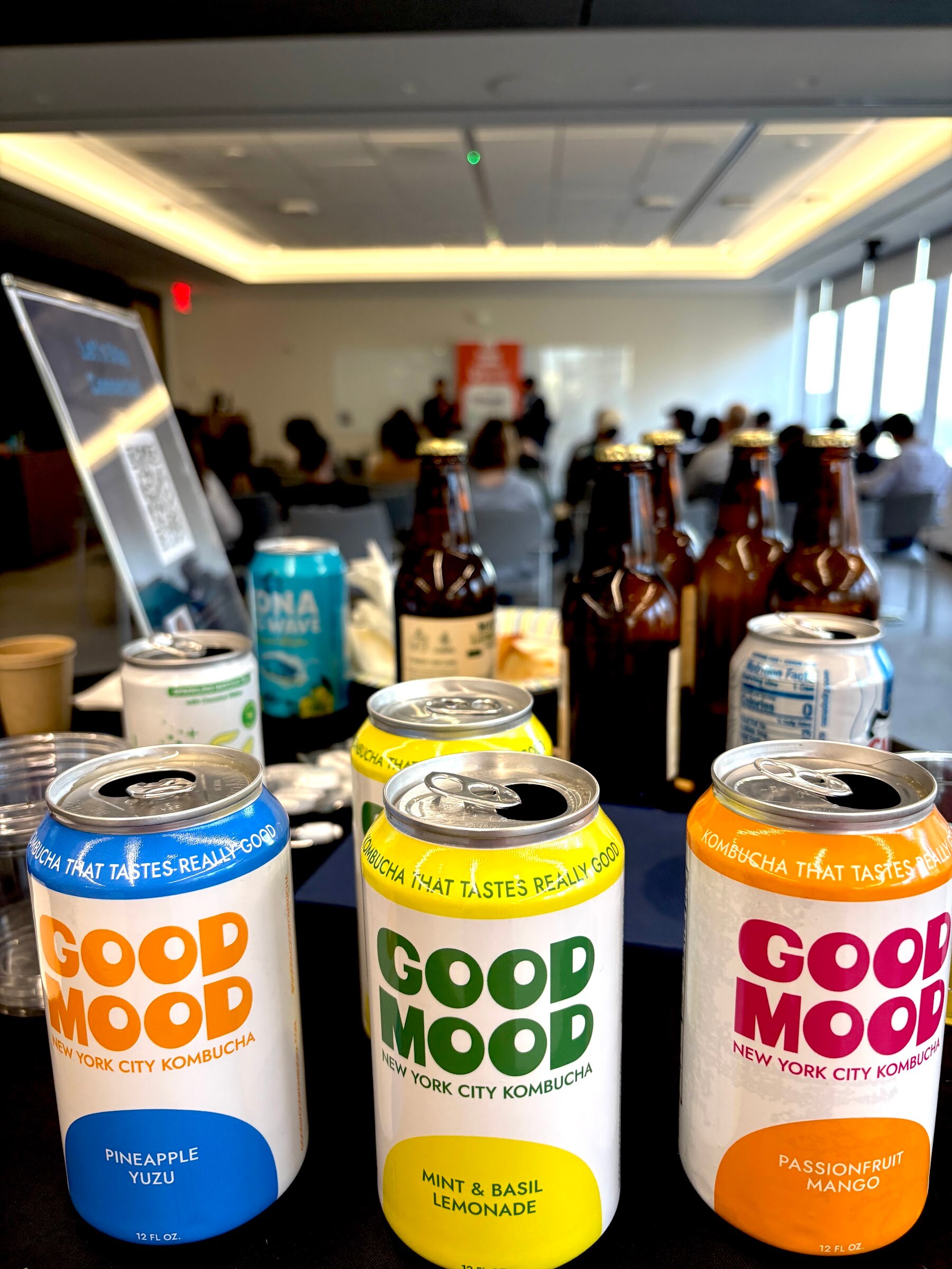 Three cans of Good Mood kombucha in front of bottles and cans on a sampling table.