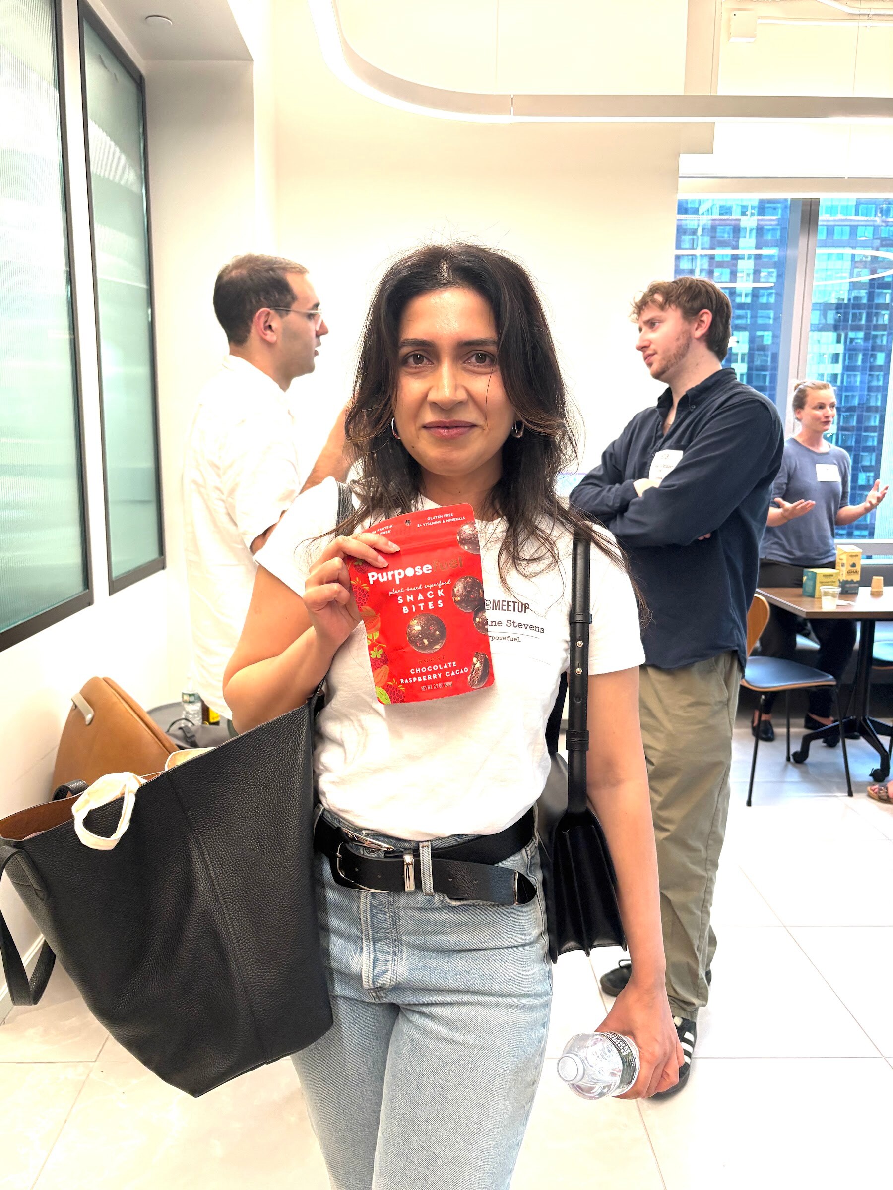 Woman with a black tote holds up a red bag of PurposeFuel snack bites in a bright room of attendees.