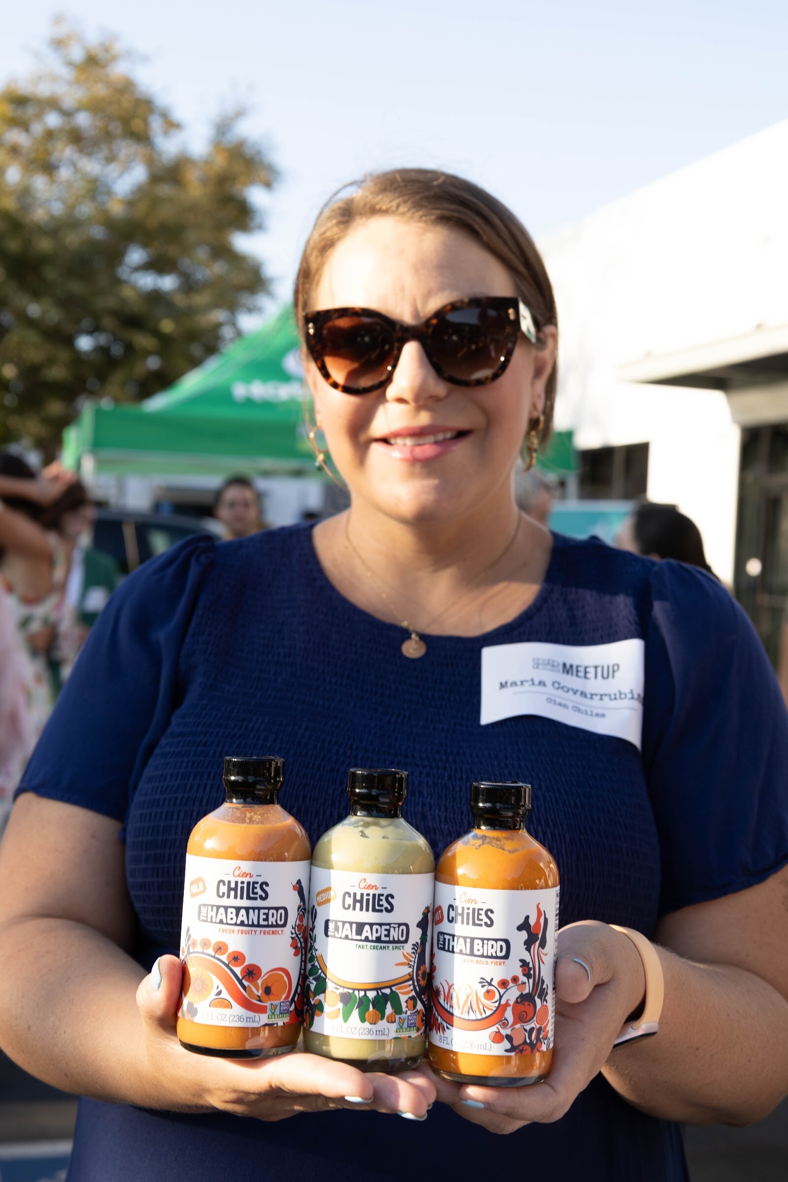 Smiling woman in sunglasses and a name badge holding three bottles of Chiles hot sauce.
