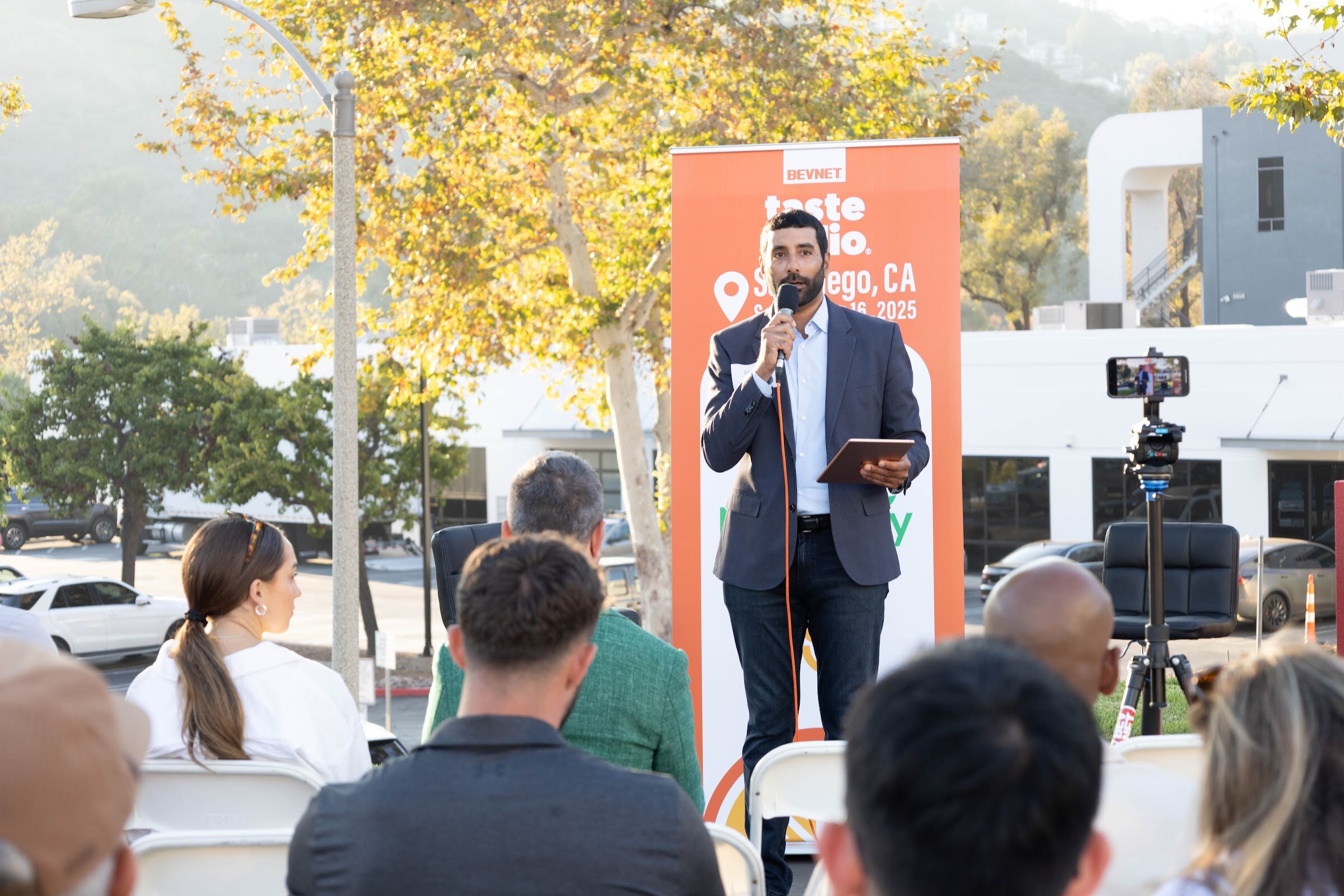Man in a blazer holding a microphone and a tablet in front of an orange Taste Radio banner, with seated guests in front.