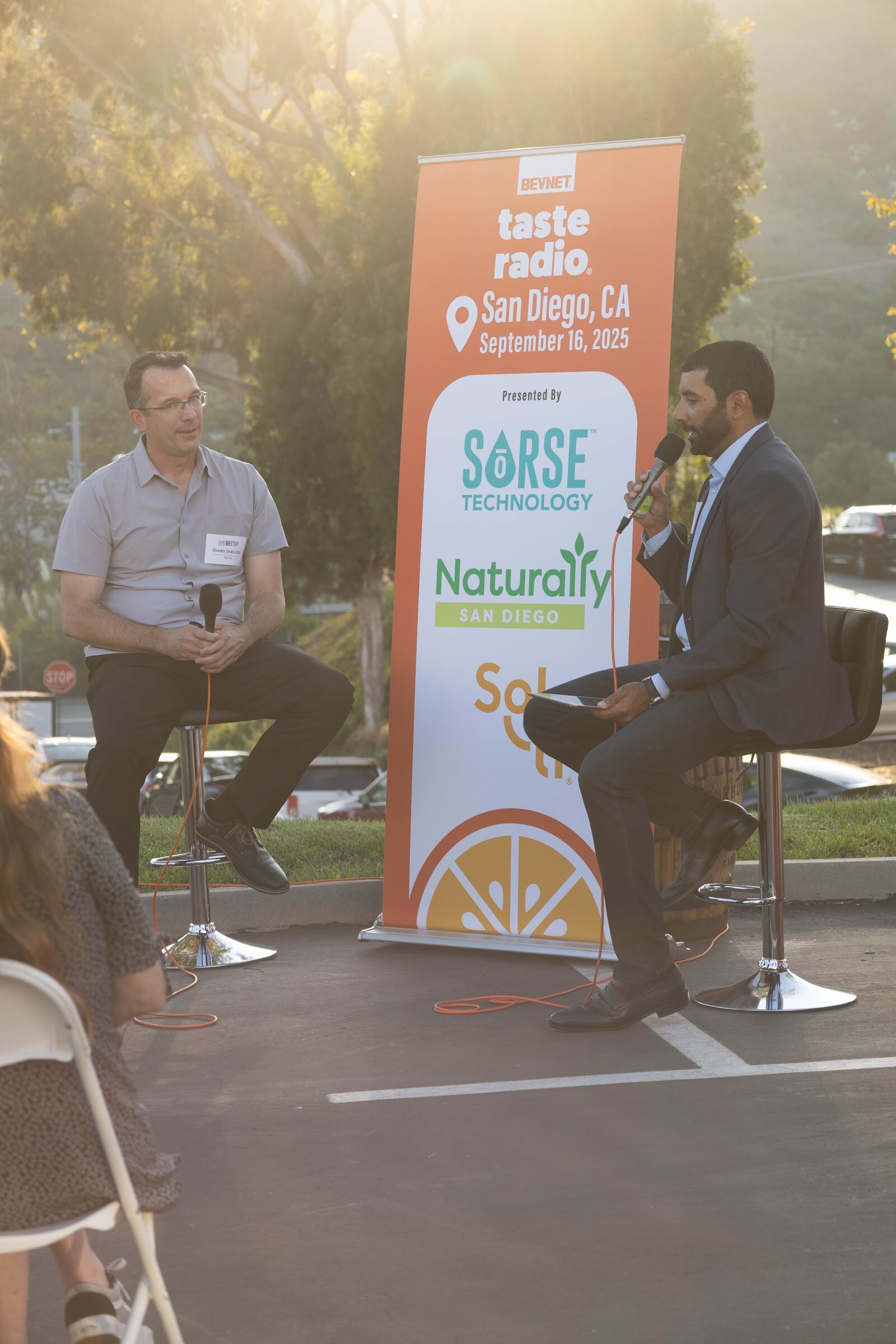 Man in a gray shirt and the host in a suit seated on stools in front of a banner listing Sorse, Naturally San Diego and Sol-ti.