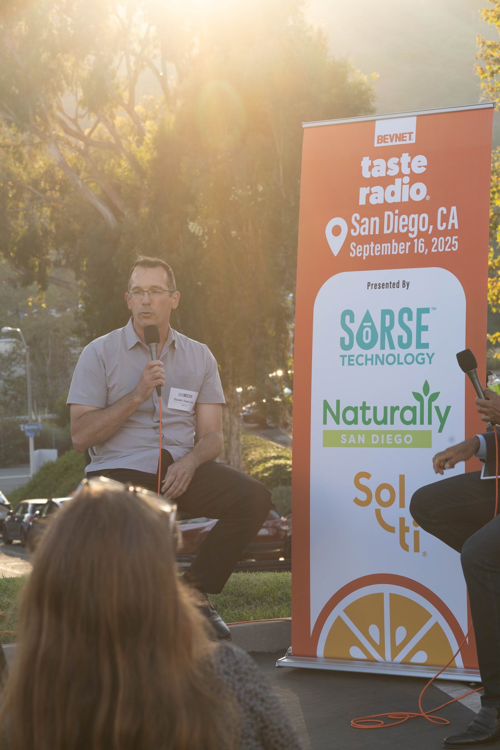 Man in a light shirt holding a microphone beside a banner for Sorse Technology and Naturally San Diego.