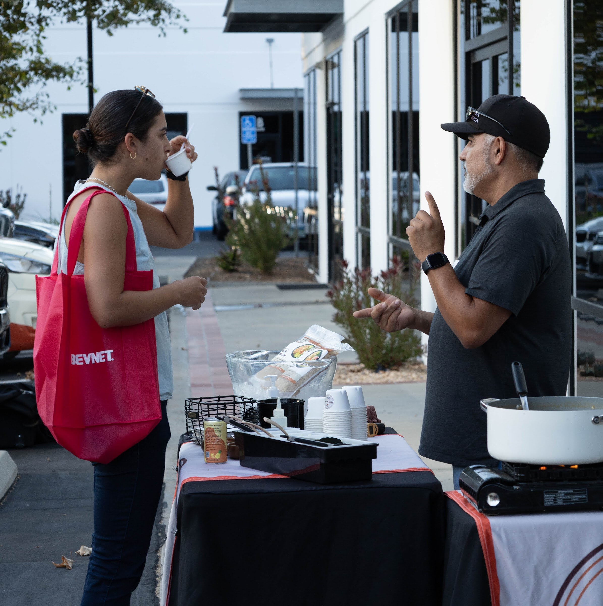 Woman in a red tote drinking from a cup beside a man in a black cap at a table with a pot and stove.