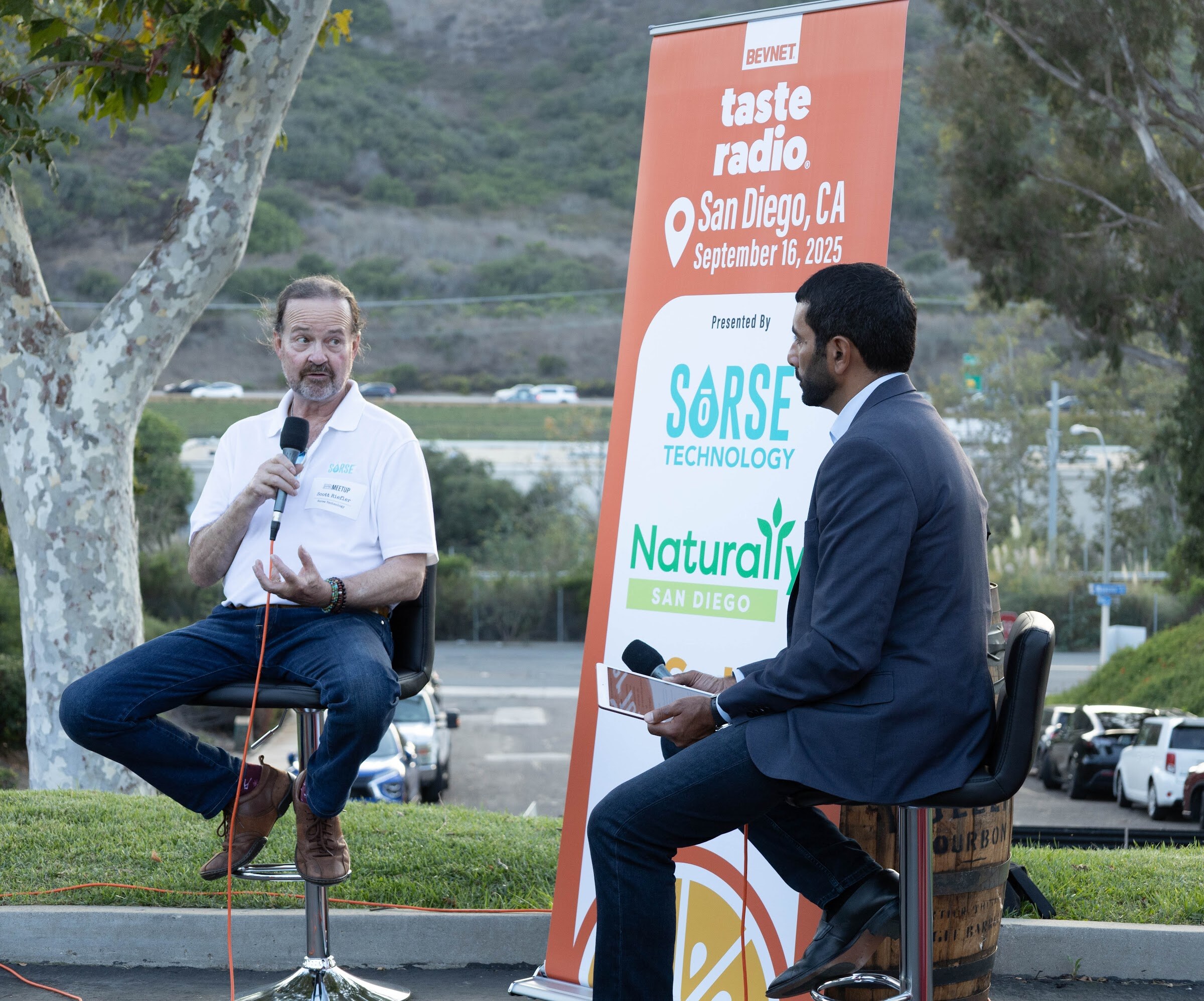 Man in a white shirt holding a microphone beside the host, in front of the Taste Radio banner.