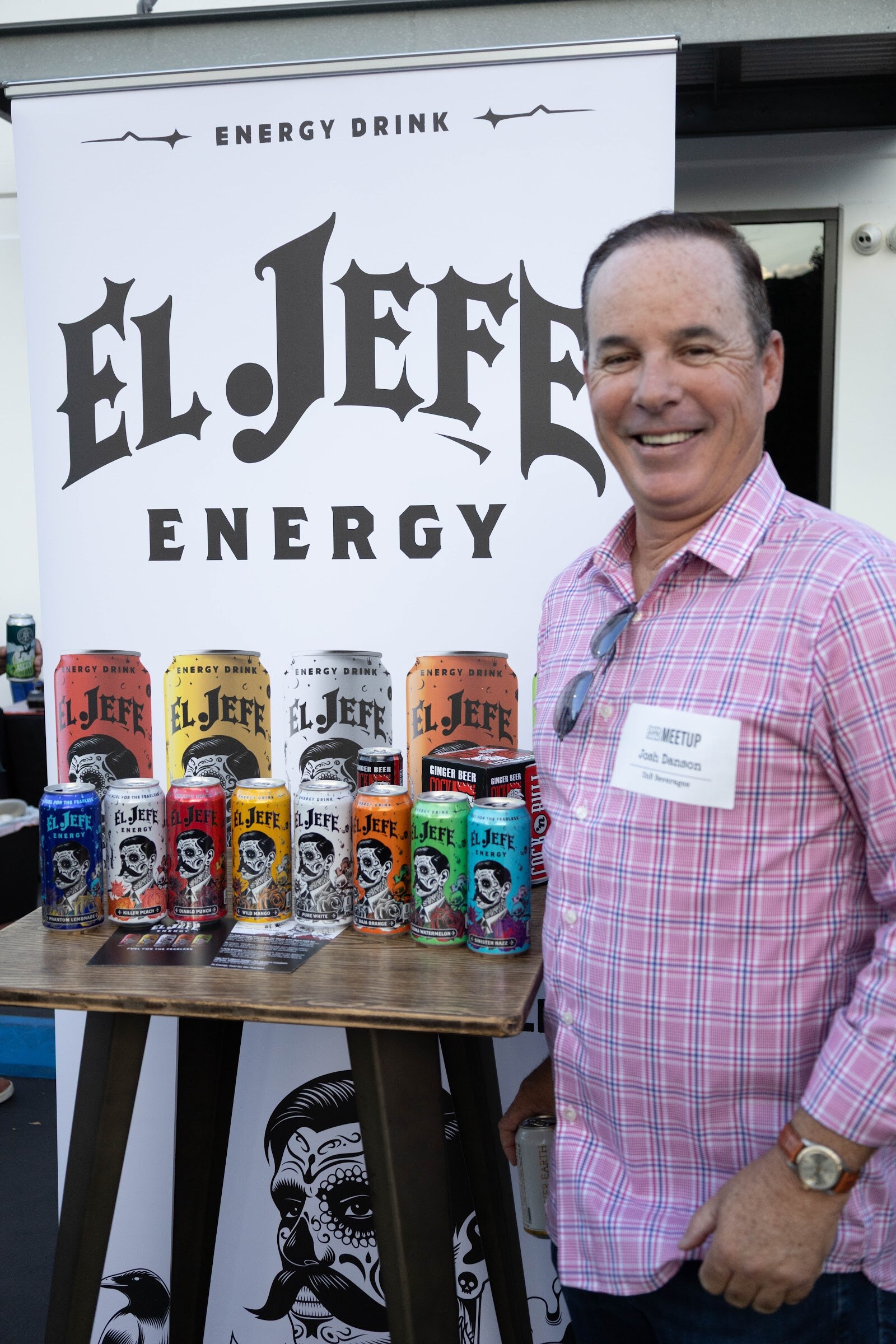 Smiling man in a pink checked shirt beside an El Jefe Energy banner and cans.