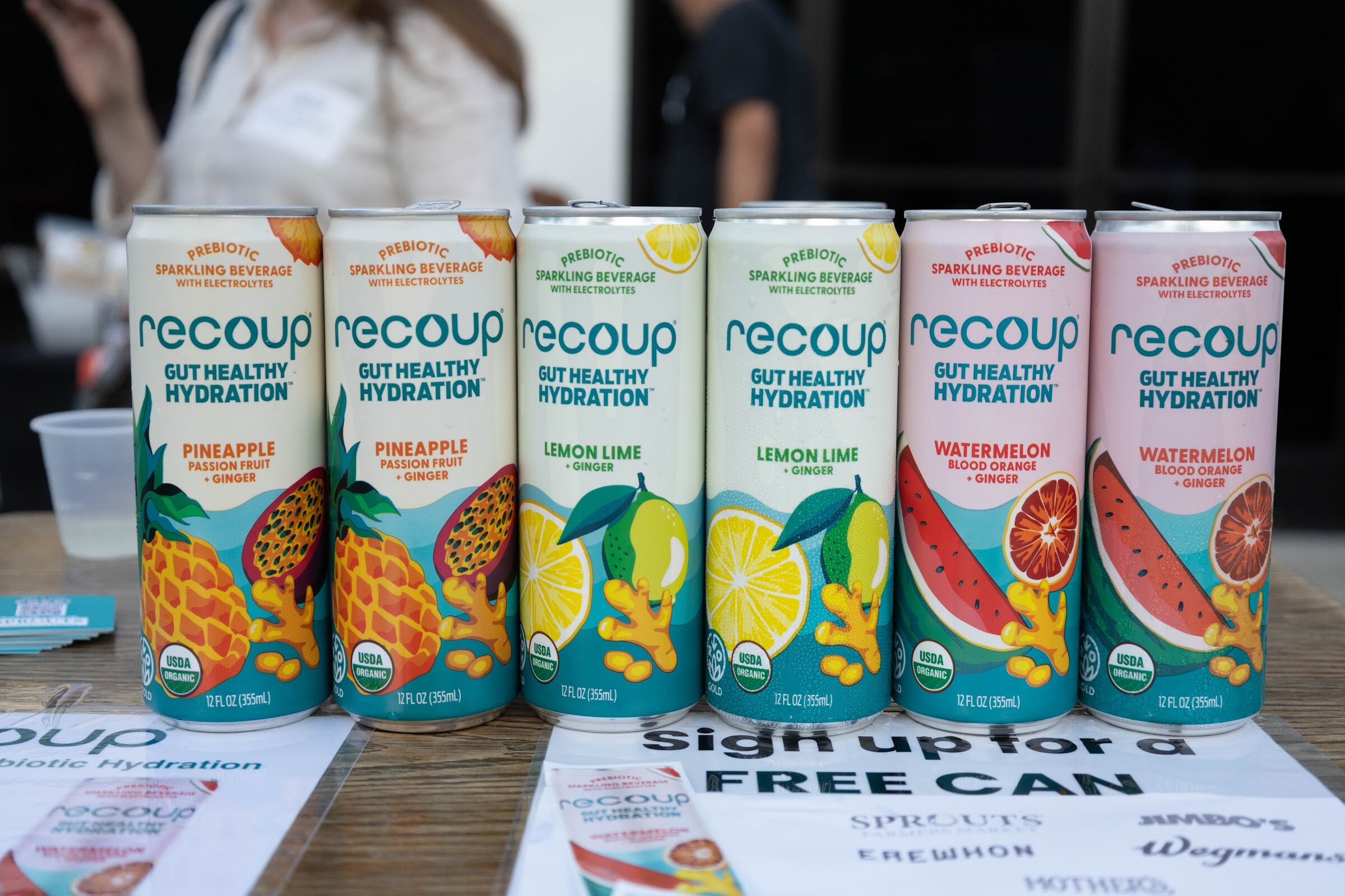 Row of Recoup cans in pineapple, lemon lime and watermelon with a sign-up sheet.