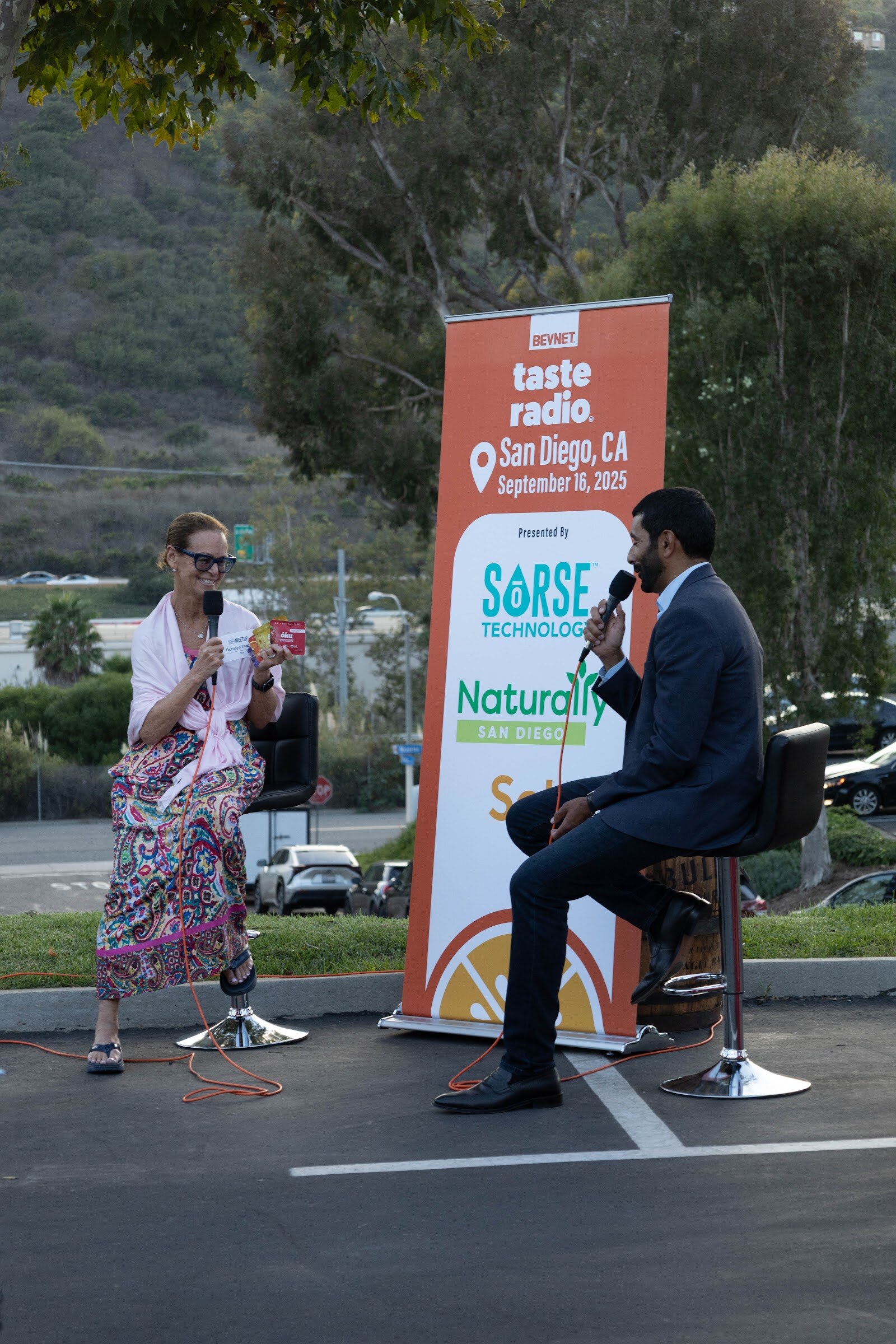 Woman in a pink floral dress holding a microphone beside the host on stools.